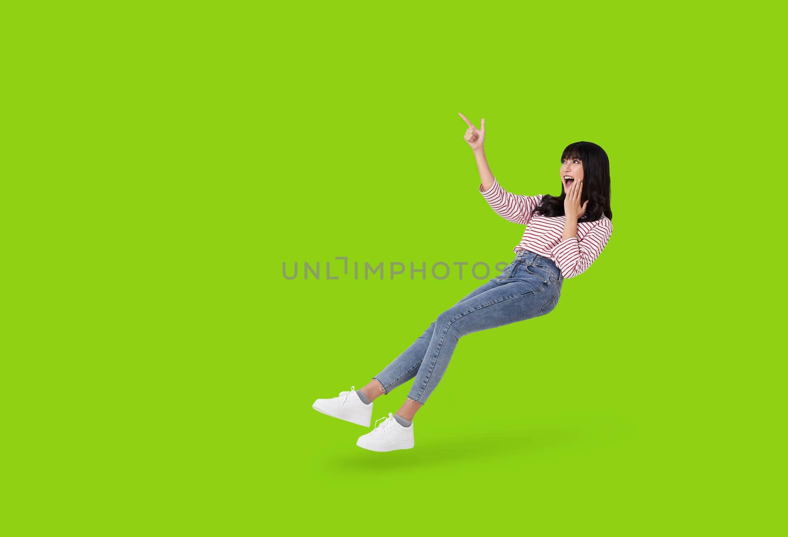 Young beautiful smiling and surprise asian girl floating in mid air with hand pointing up to copy space isolated on green background. by barameeyay