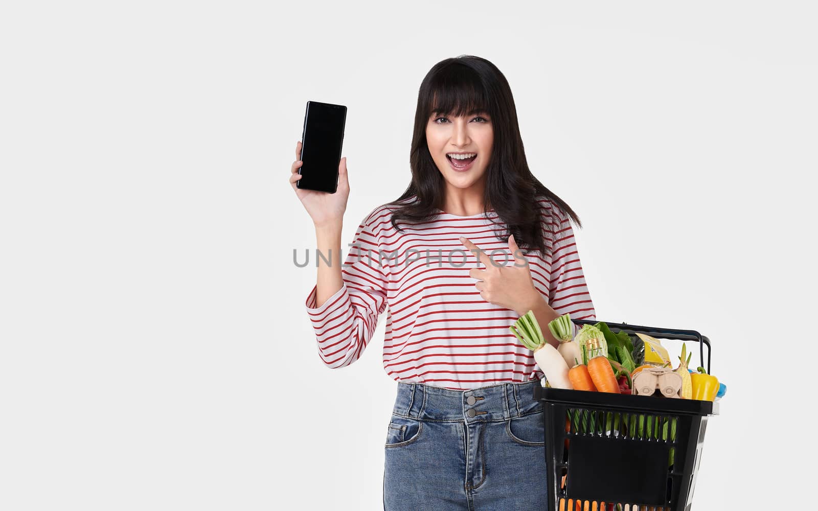 Happy Asian woman with smartphone holding basket full of fresh vegetable groceries isolated on white background.