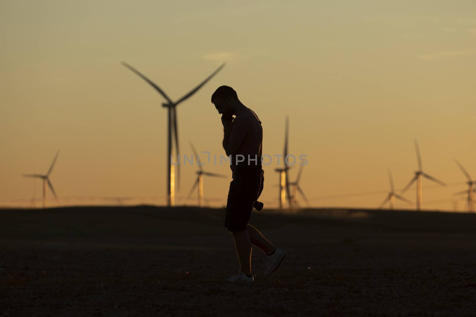A man walks talking on the phone in the countryside at sunset, after taking pictures of distant wind turbines, on the horizon.