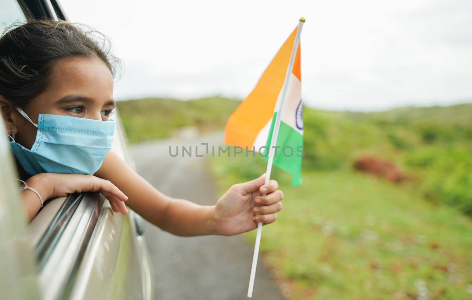 Young girl kid with medical mask holding Indian flag in moving car window - Concept of celebrating Independence or republic day during coronavirus or covid-19 pandemic.