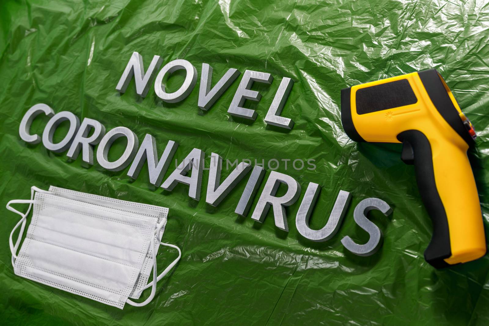 words novel coronavirus laid with metal letters on green crumpled plastic film backdrop with face masks and infrared thermometer - diagonal perspective with dramatic light