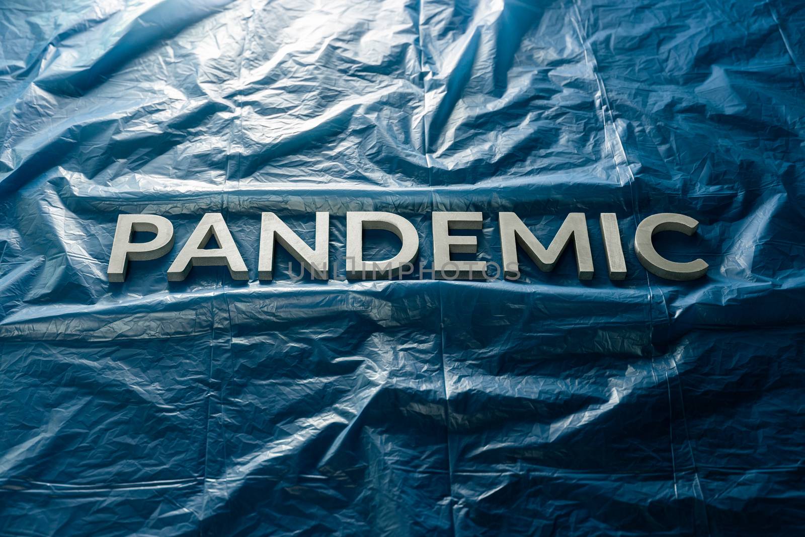 The word pandemic laid with silver letters on crumpled blue plastic film background Slanted centered with dramatic cold light.