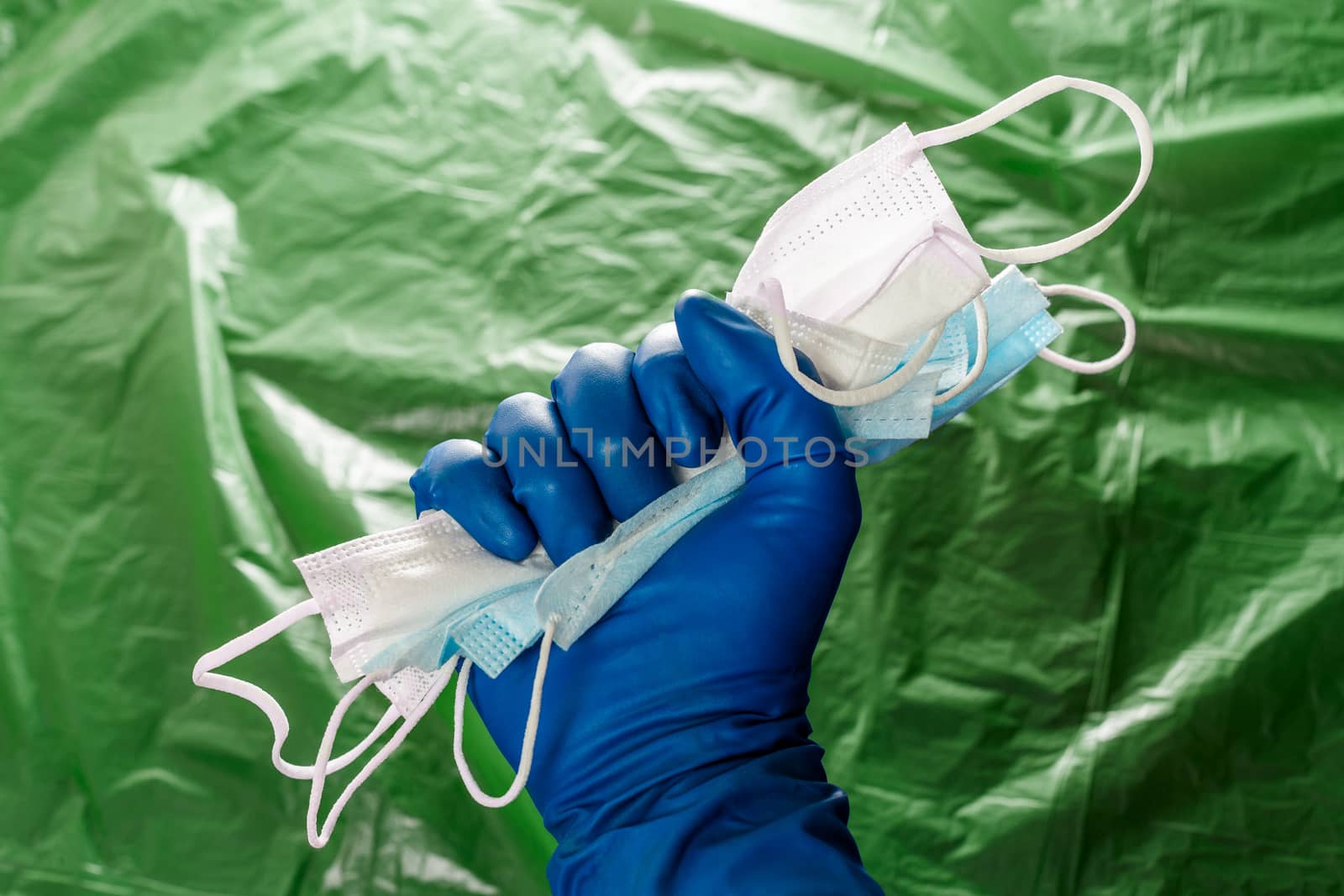 A hand in blue medical glove holding a bunch of face masks on blurry green plastic film background. Symbol of coronavirus pandemic resistance and help.