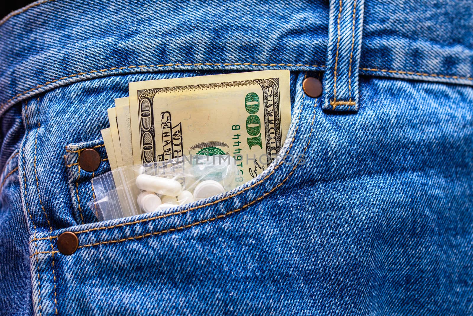 White drug pills and US dollar banknotes in the front left pocket of blue jeans. Concept of medical expenses or illegal drug dealing