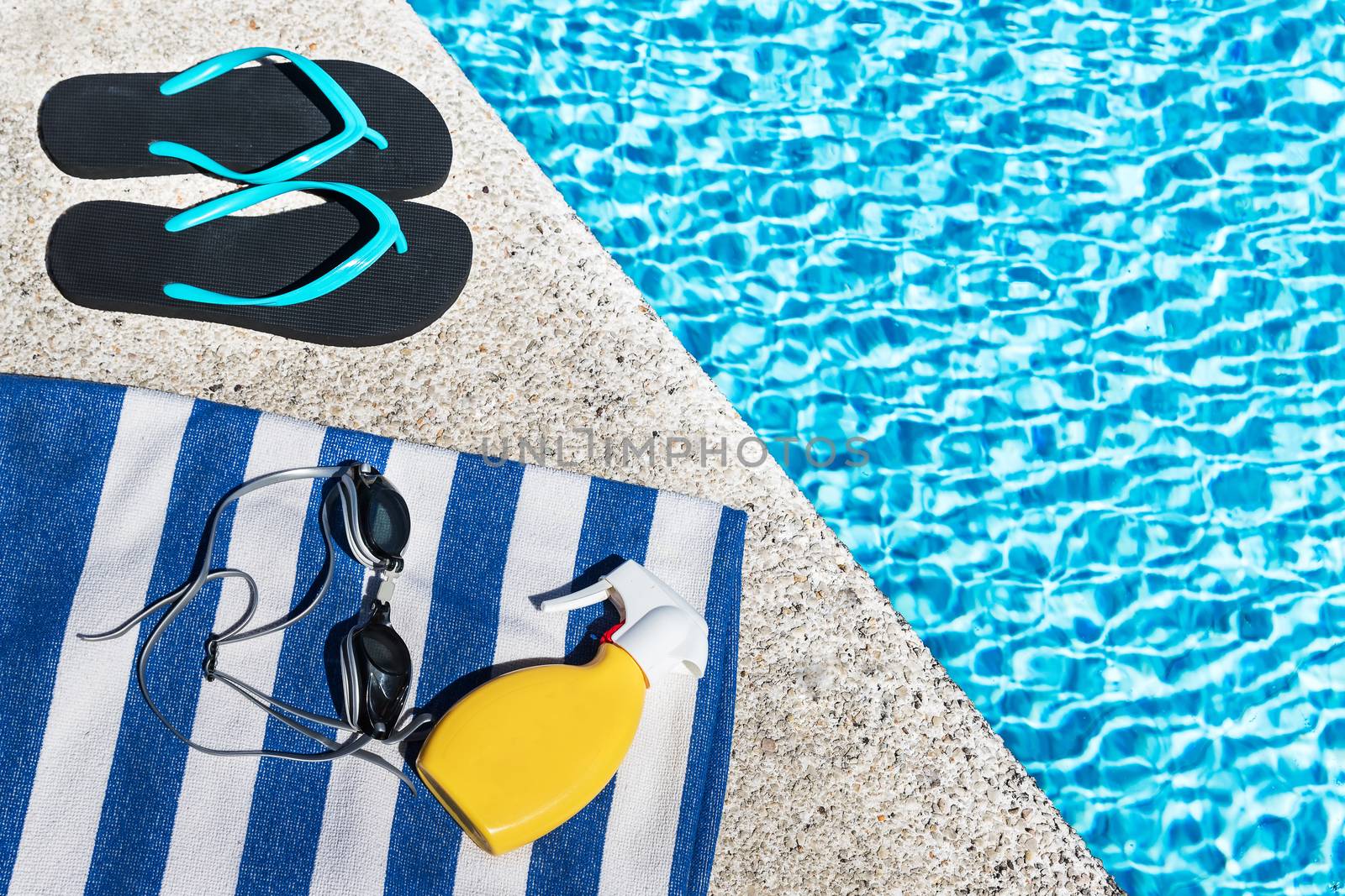 Flip flops, sunscreen spray and swimming goggles on a blue and white striped towel at the edge of a pool.. Copy space. Top view.