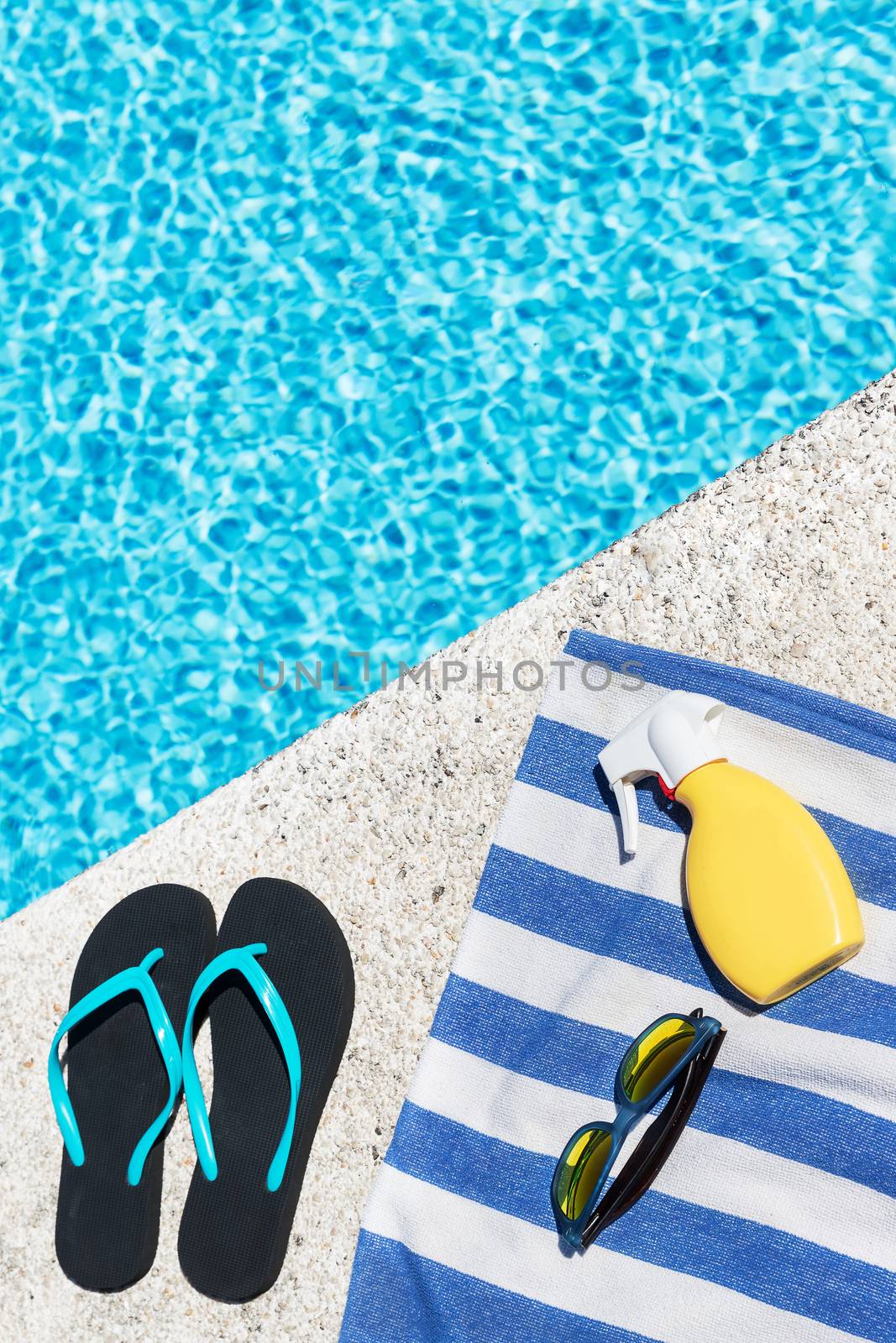 Flip flops, sunscreen spray and sunglasses on a blue and white striped towel at the edge of a pool. Copy space. Top view.