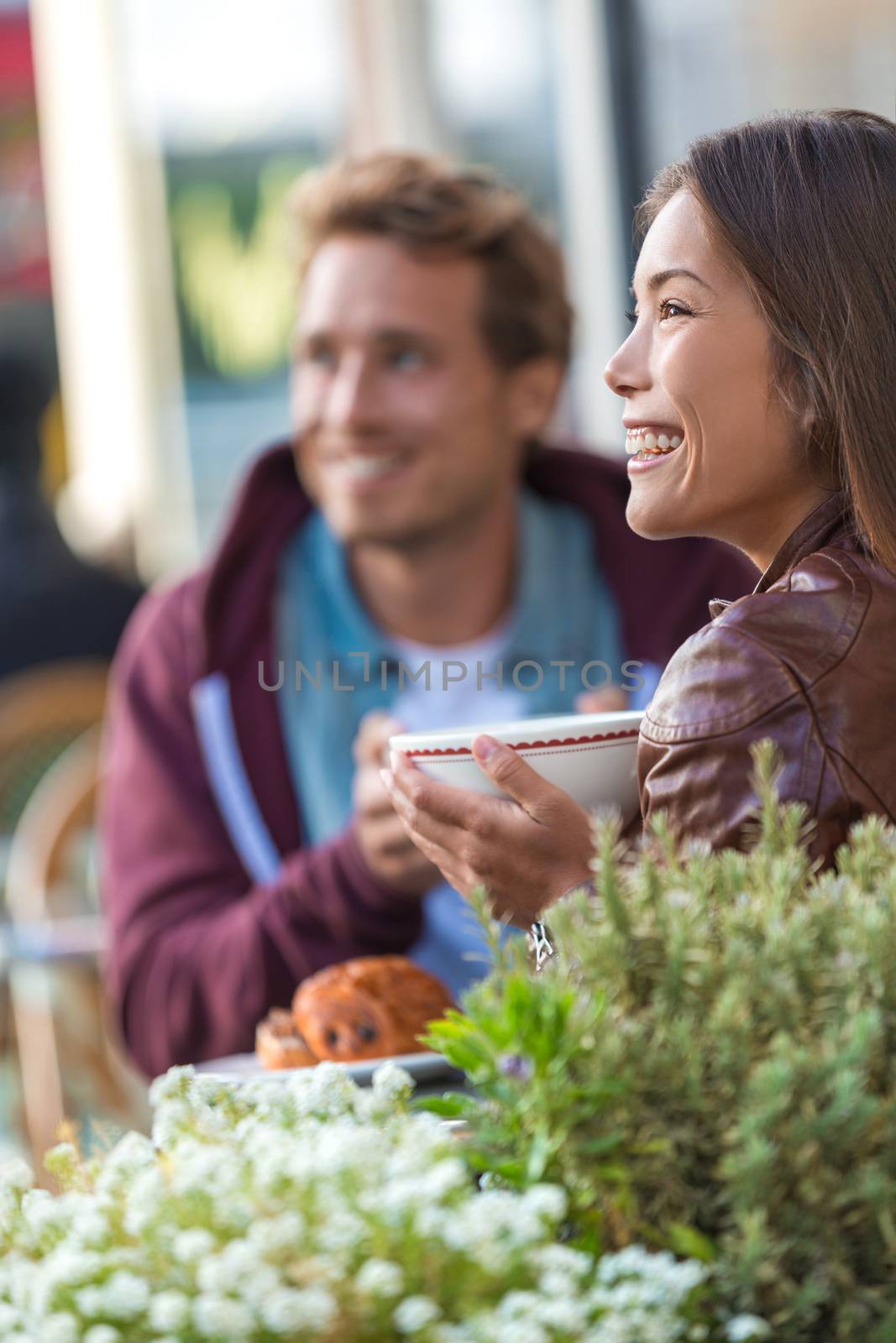 Happy people eating brunch at cafe. Young couple hipsters drinking coffee at restaurant table outside sidewalk terrace at parisian bistro in european city. Asian woman happy with boyfriend or friend. by Maridav