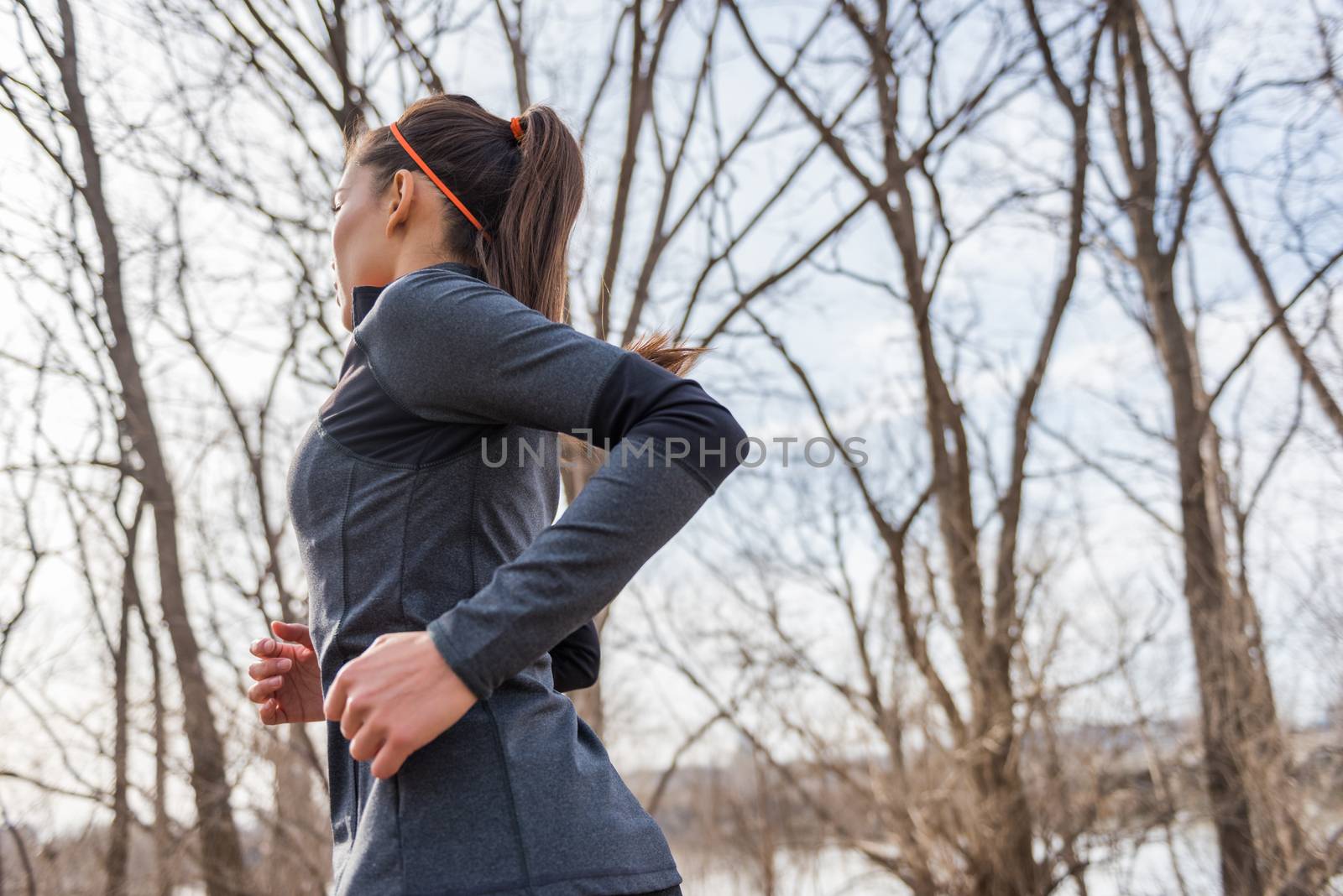 Young fitness woman runner running on trail. Girl athlete working out cardio in autumn or winter nature outdoors on forest trail in sunlight fall wearing cold weather jacket.