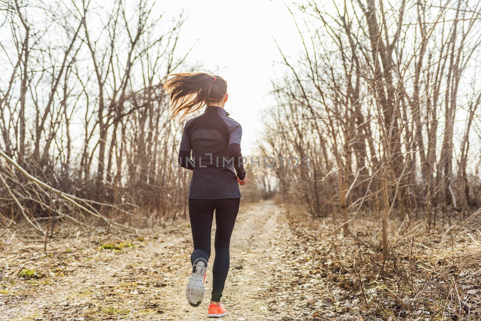 female athlete runner running in autumn nature outdoors on forest trail path in beautiful sunny sunlight fall cold weather. Unrecognizable woman in activewear tights jogging away view from behind.