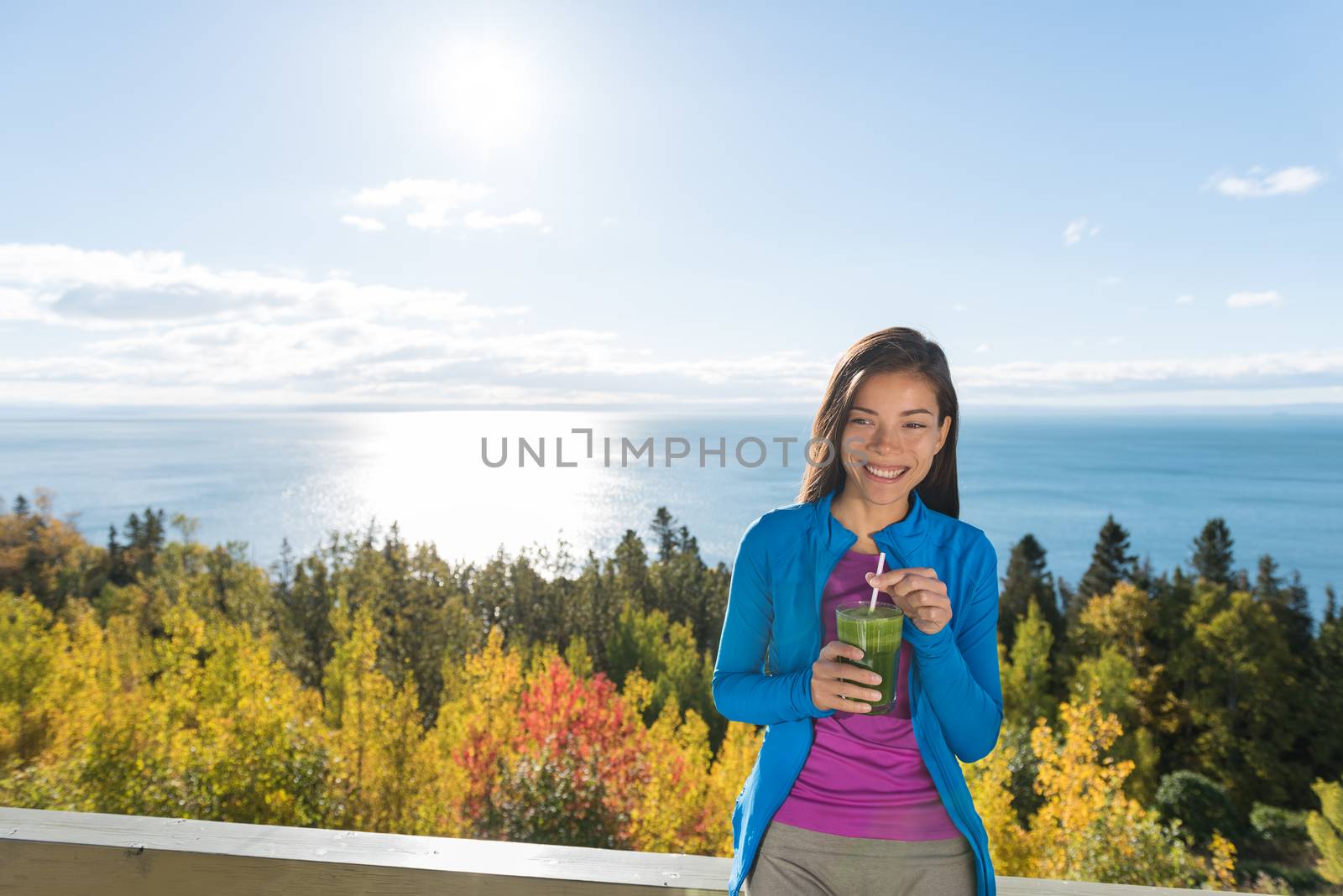 Outdoor fall nature asian woman drinking fresh green vegetable smoothie as a healthy breakfast or lunch detox on home terrace in beautiful morning nature outdoors. Happy smiling fitness girl.