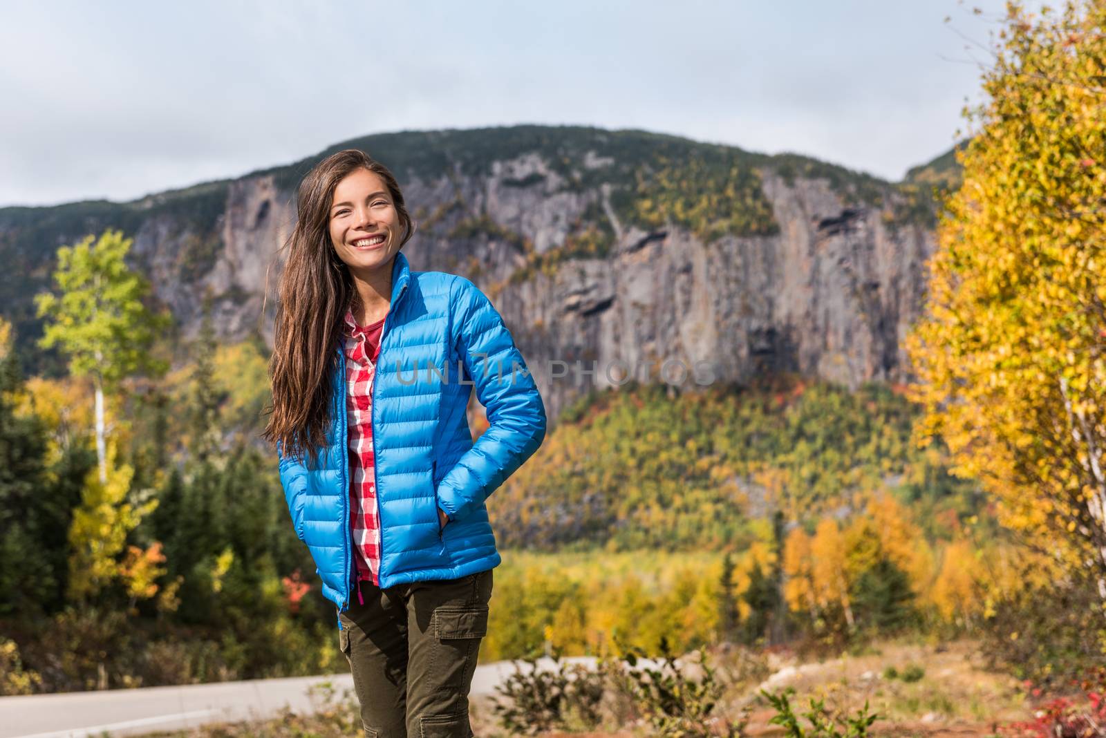 Autumn nature lifestyle happy asian woman relaxing outdoors on camping travel park with mountain background. Hiking girl wearing blue down jacket enjoying fall season scenic landscape.