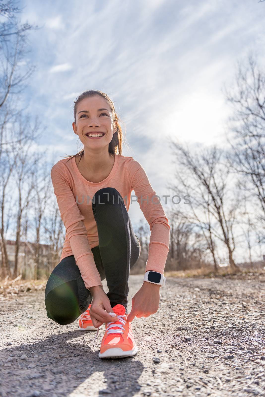Autumn trail runner asian girl tying running shoes wearing sports smartwatch gadget gear. Female active athlete lacing shoe laces on forest path using smart watch heart rate fitness monitor.