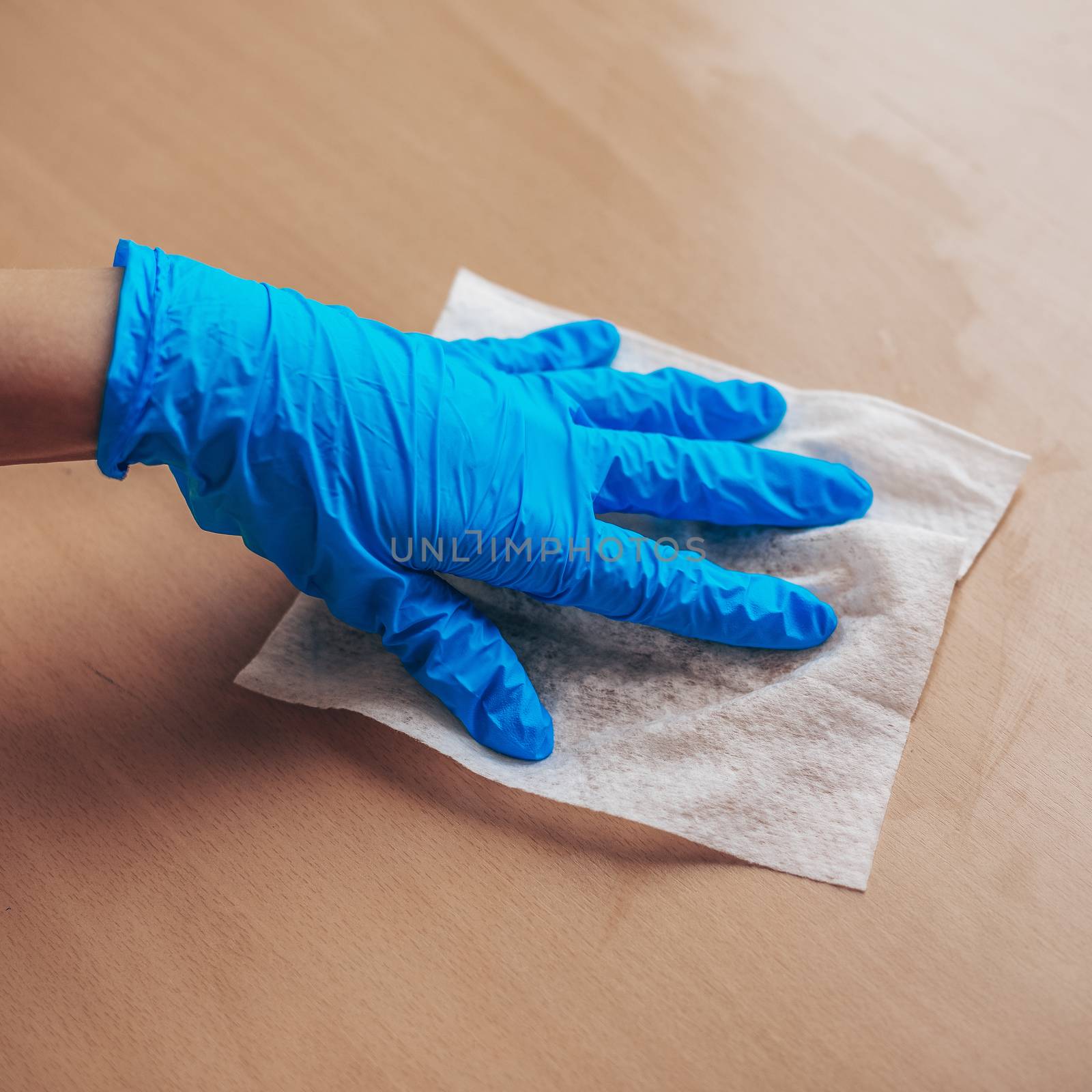 Woman's hand in blue gloves sanitizing cleaning home office wood table surface with wet wipes