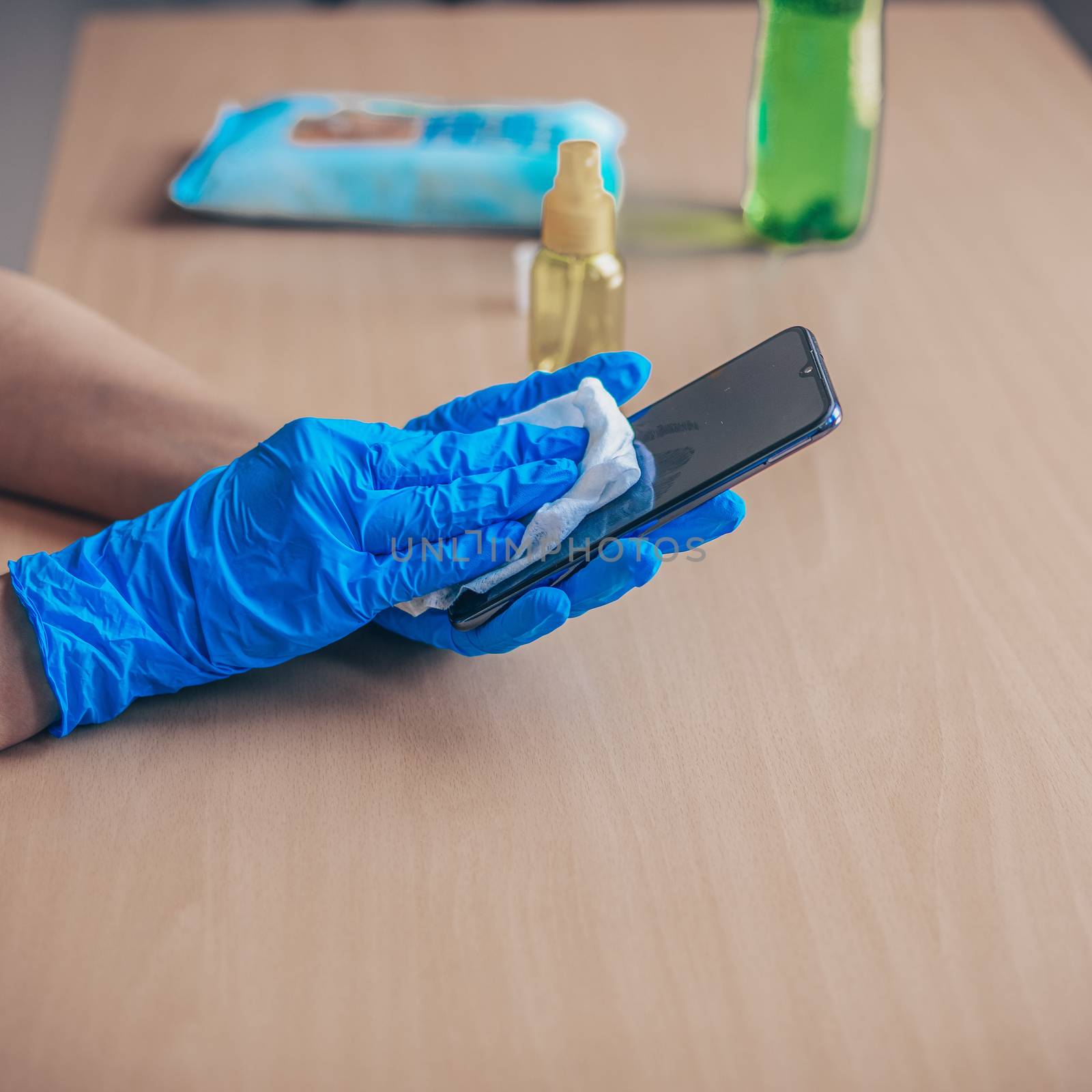 Woman's hand in blue gloves sanitizing cleaning smartphone mobile phone on wood table surface with wet wipes and alcohol