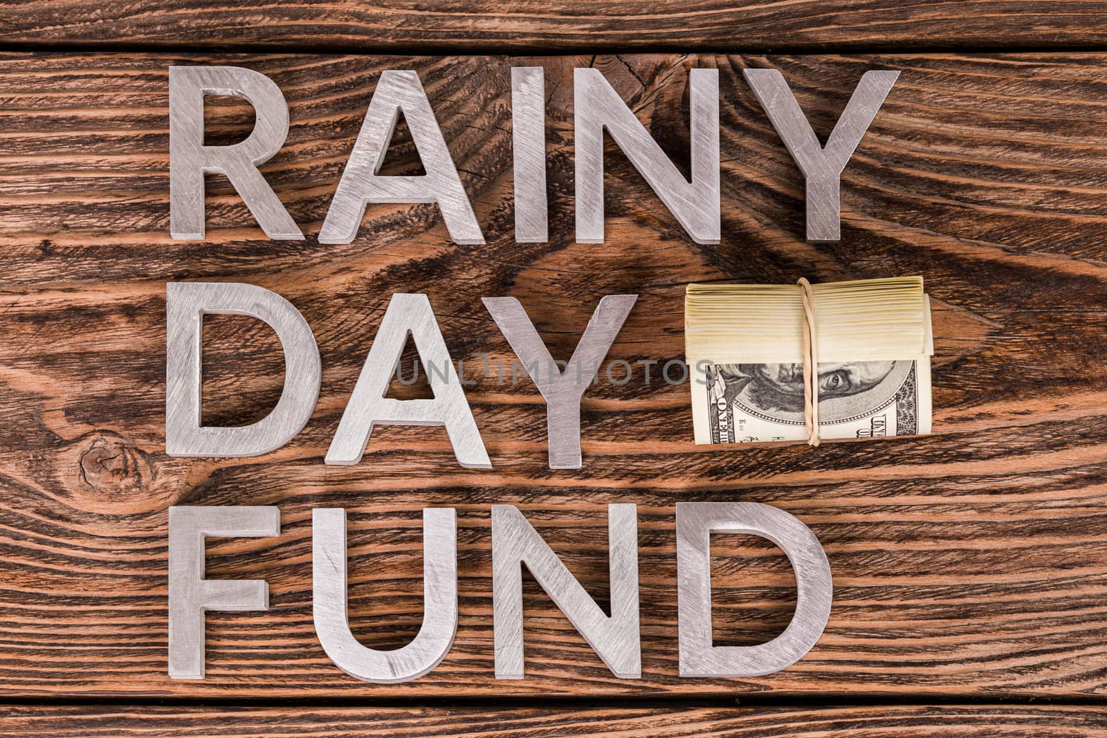 words RAINY DAY FUND laid on wooden surface by metal letters with small roll of us dollar banknotes - flat lay above view