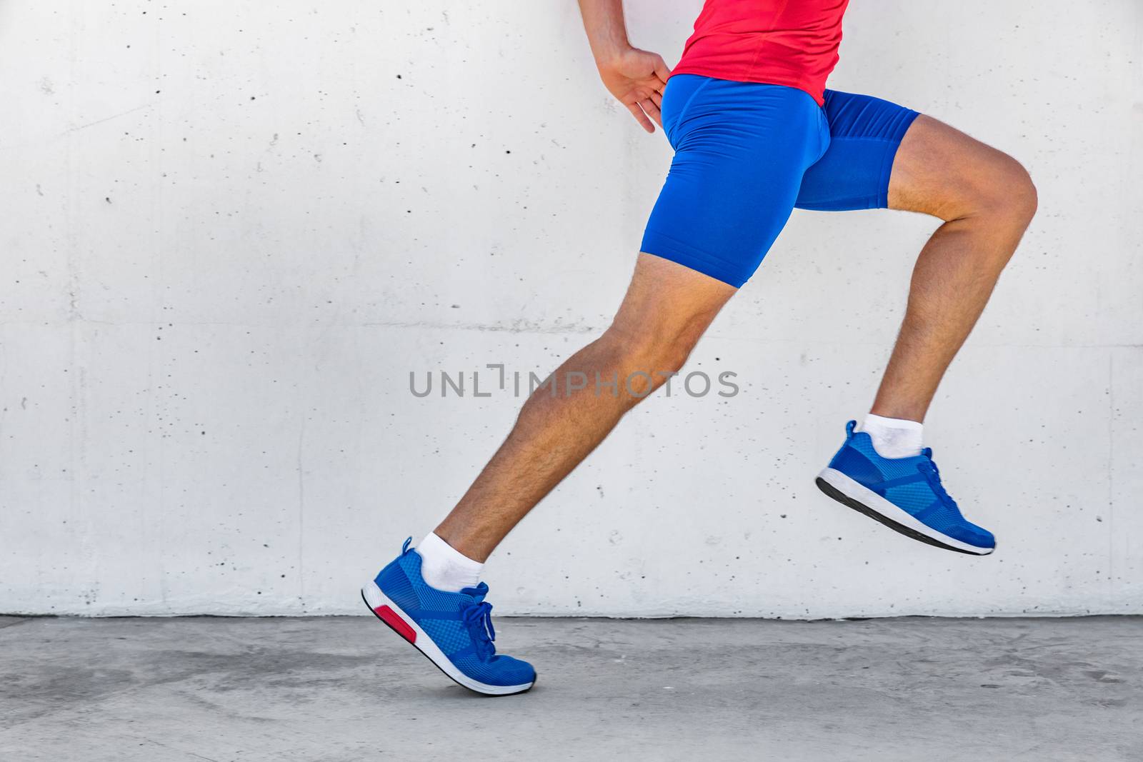 Fitness athlete man running on asphalt sidewalk in city street. Sport lifestyle. Closeup of lower body runner's legs on workout marathon run against white wall background. Shoes, compression clothing.