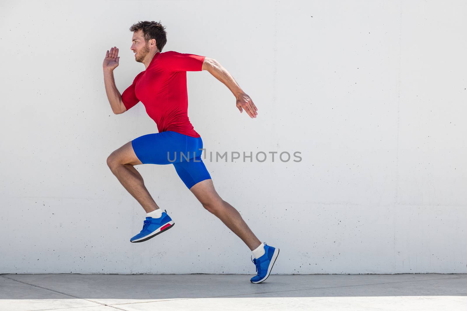 Running man runner training doing outdoor city run sprinting along wall background. Urban healthy active lifestyle. Male athlete doing sprint hiit high intensity interval training.