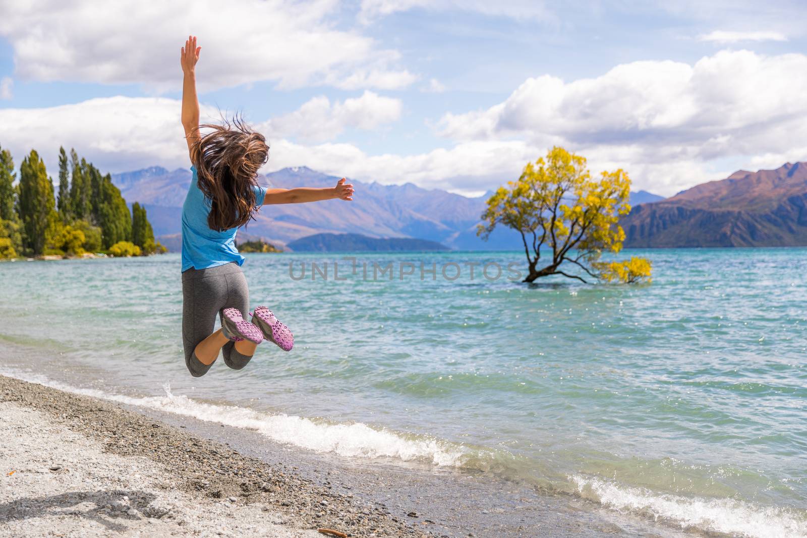New Zealand travel happy tourist woman jumping of joy at Wanaka lake landscape with lone tree, famous attraction. by Maridav