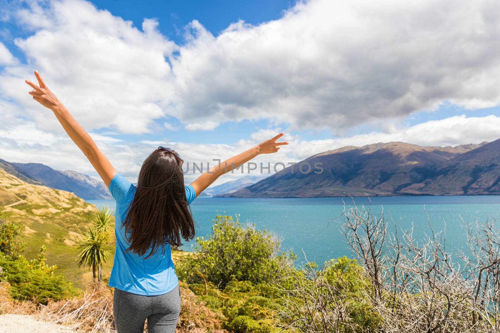New Zealand travel happy tourist woman with arms up at Wanaka lake nature landscape outdoors. Wanderlust adventure young girl with peace hand sign. by Maridav