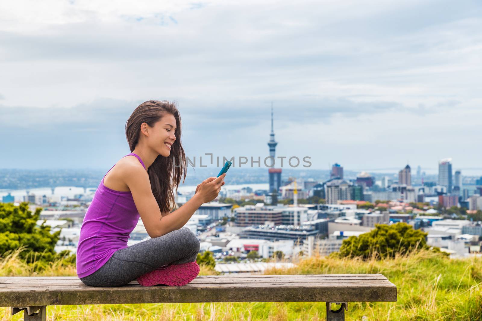 Auckland city runner girl using mobile phone app at skyline summer park. View of cityscape from Mount Eden, in North Island, New Zealand. Girl taking break outdoors after running.