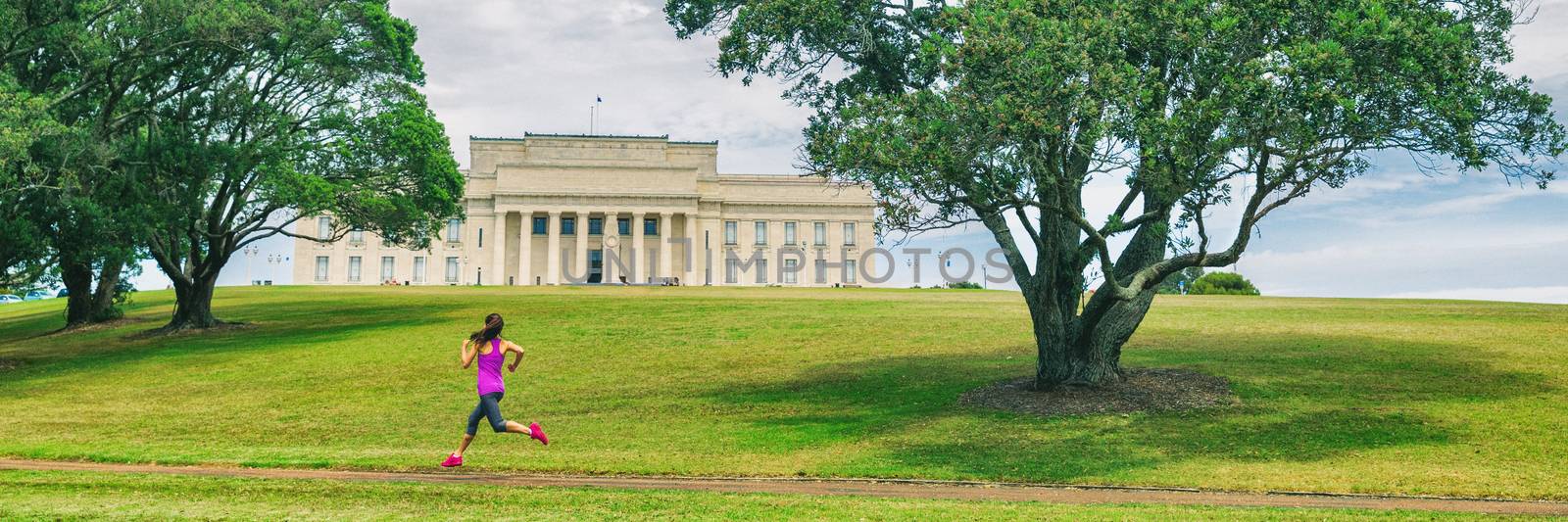 Park runner woman running in Auckland city banner. Panoramic crop of jogging girl on green grass and trees at Auckland Domain park with Memorial museum background. Urban lifestyle. by Maridav