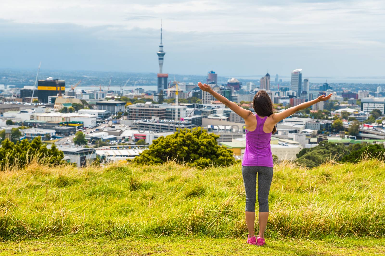 Auckland city skyline view from Mount Eden of Sky tower, New Zealand. Happy woman with arms up in freedom and happiness at top of Mt Eden urban park famous tourist attraction. by Maridav