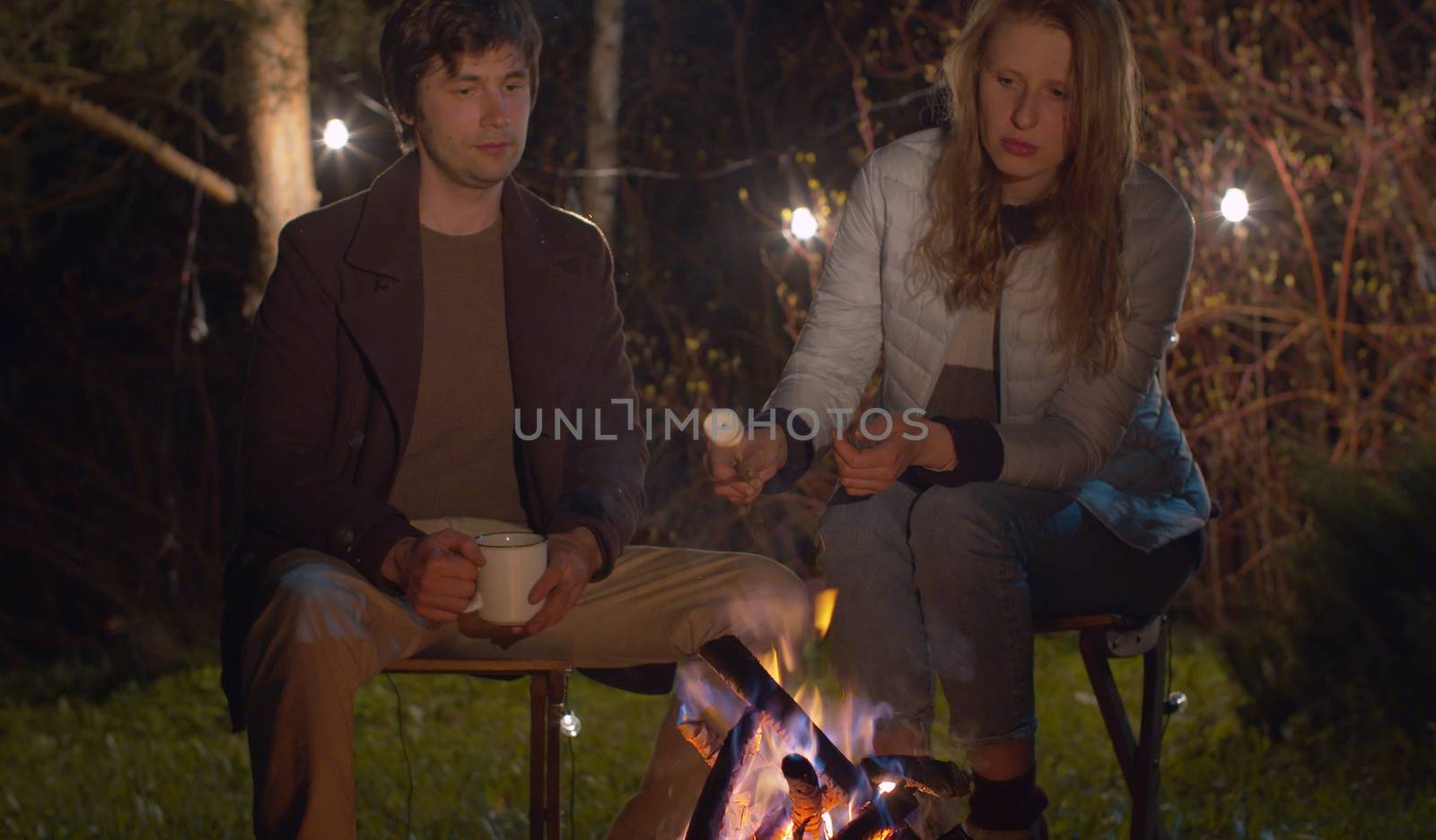 Young happy couple near the bonfire at night in the forest. Family on weekend. Man drinking tea, woman frying marshmallow. Healthy lifestyle concept