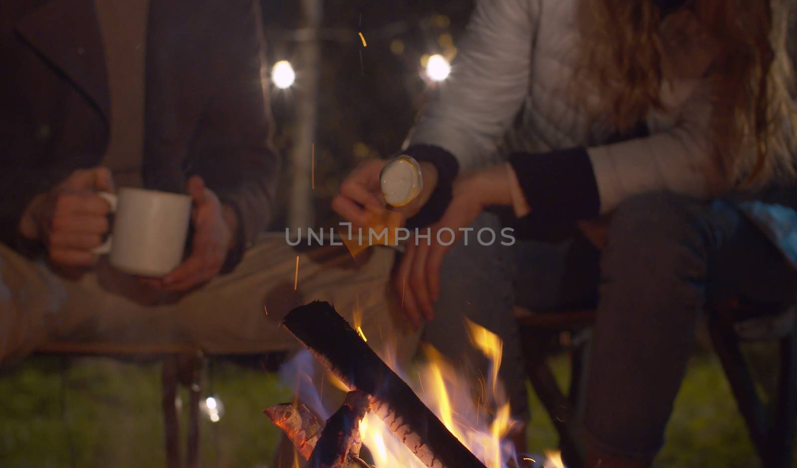 Close up female hands frying marshmallow. Young happy couple near the bonfire at night in the forest. Family on weekend. Man drinking tea. Healthy lifestyle concept