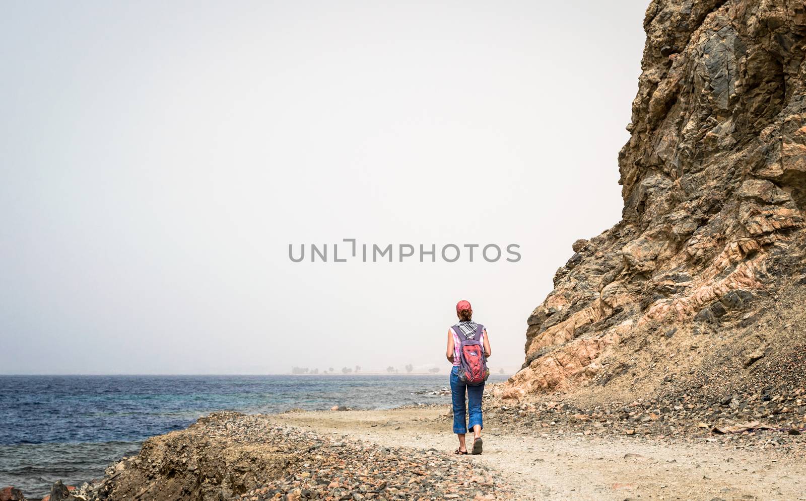 young girl tourist in a red cap with a backpack goes along the rocky shore of the Red Sea