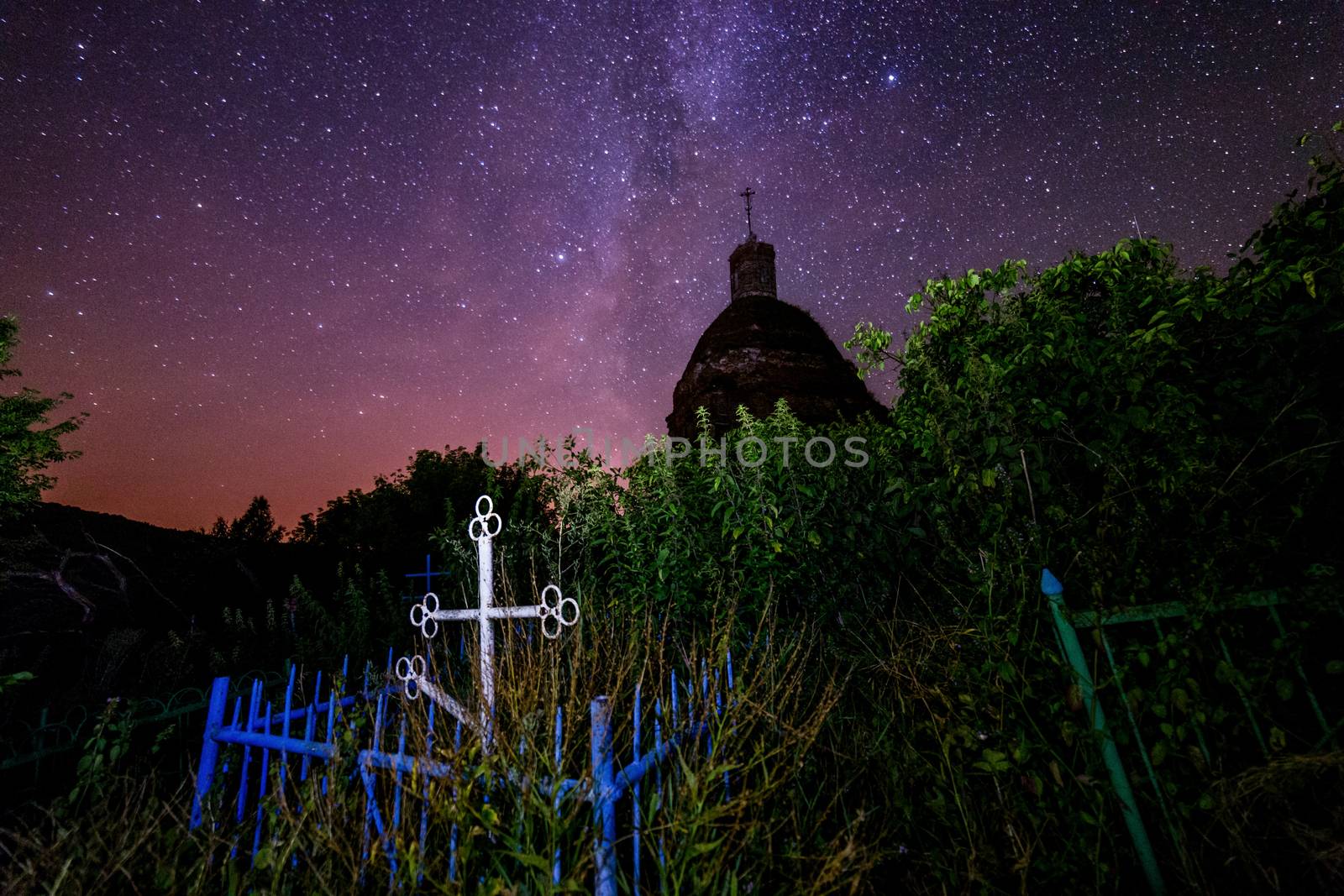 Abandoned christian grave under ruins of church at starry night with visible milky way galaxy. Tall grass and dark bushes selectively focused and lighted. No montage - real photography.
