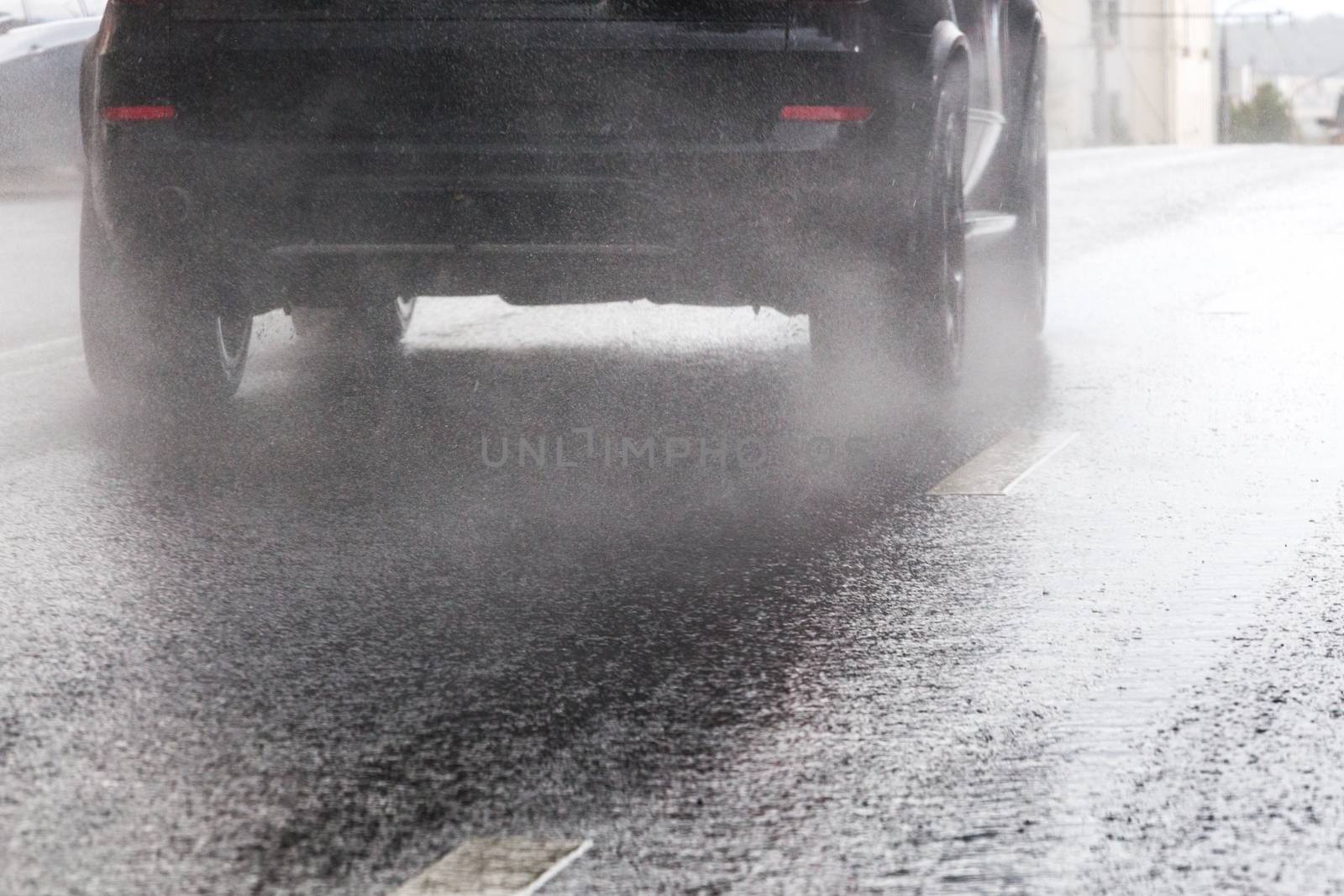 Rain water splash flow from wheels of black car moving fast in daylight city with selective focus. Tiny drops mist.