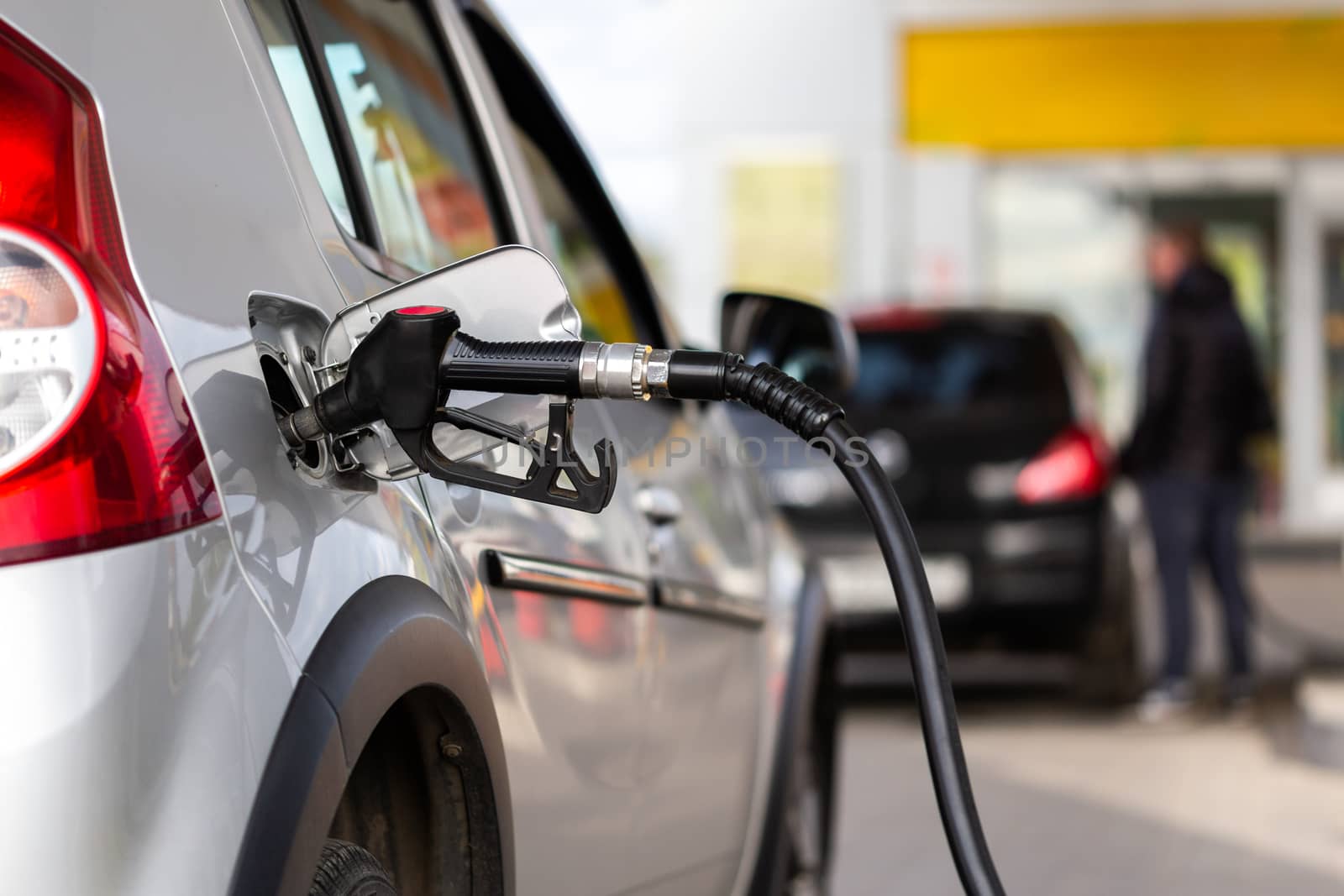 gray metallic car refueling on gas station - closeup with selective focus and blurry man on background.