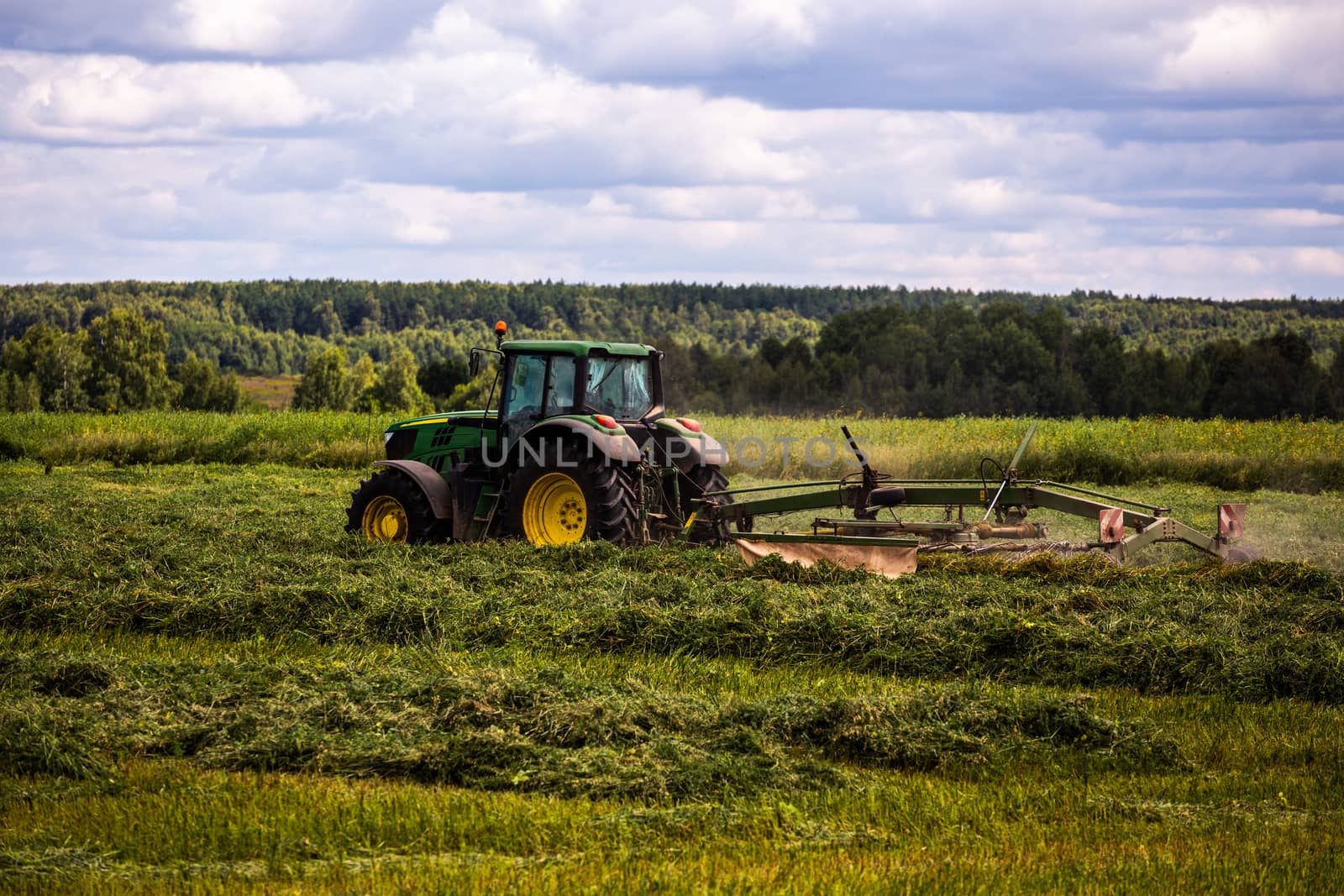 green haymaking tractor on summer field before storm - telephoto shot with selective focus and blur.