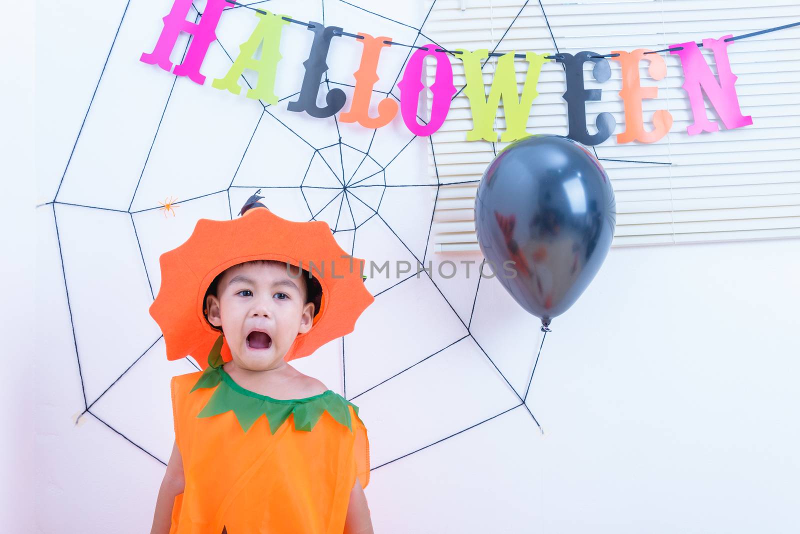 Funny happy little cute boy kid in Halloween costume with orange pumpkin Jack with Cobweb and black balloon in-home on white wall background, Happy Halloween day concept