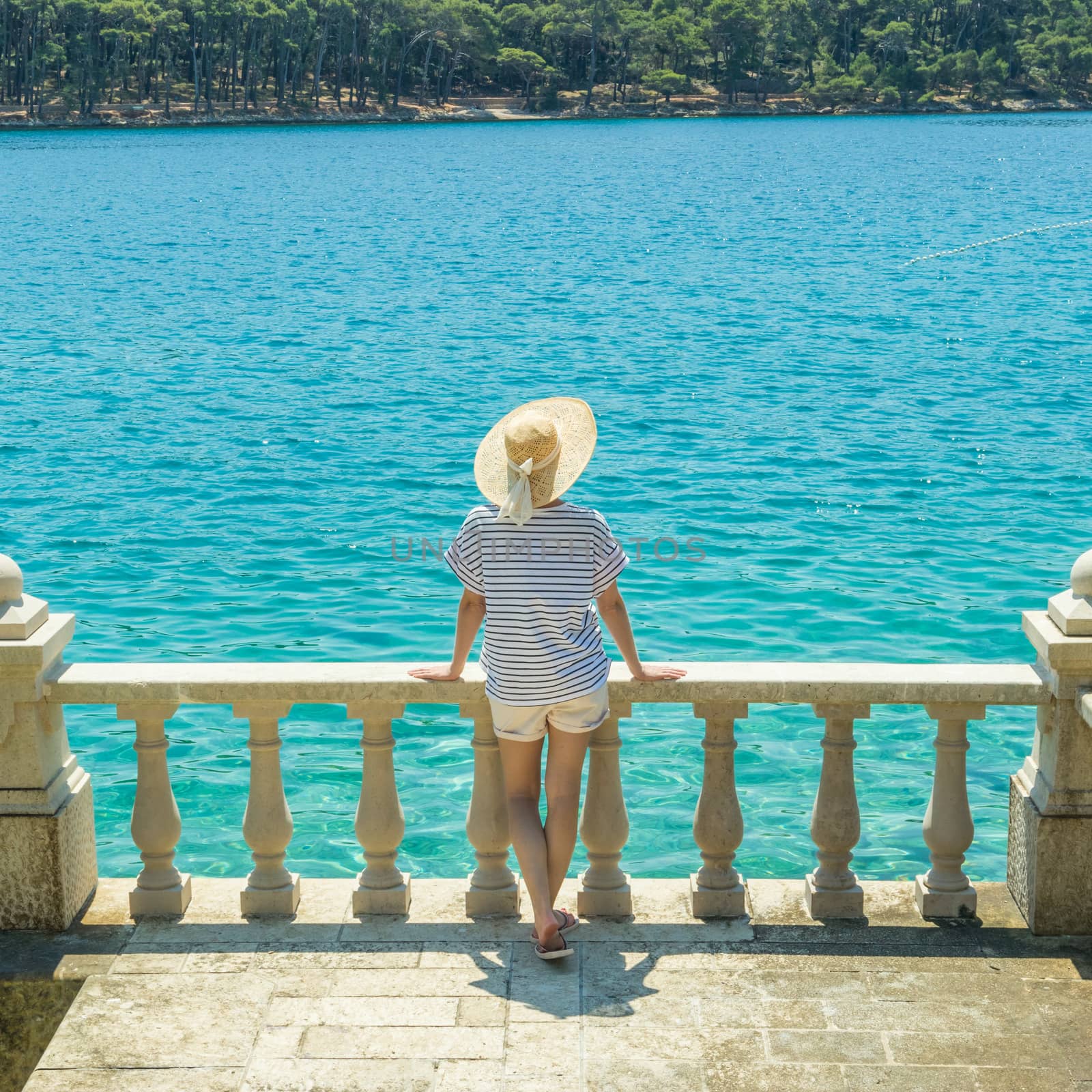 Rear view of woman wearing straw summer hat ,leaning against elegant old stone fence of coastal villa, relaxing while looking at blue Adriatic sea, on Losinj island Croatia. by kasto