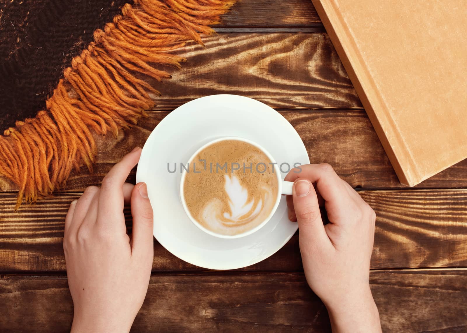 Hands holding cap of coffee on a small plate, book and wool scarf on wooden background