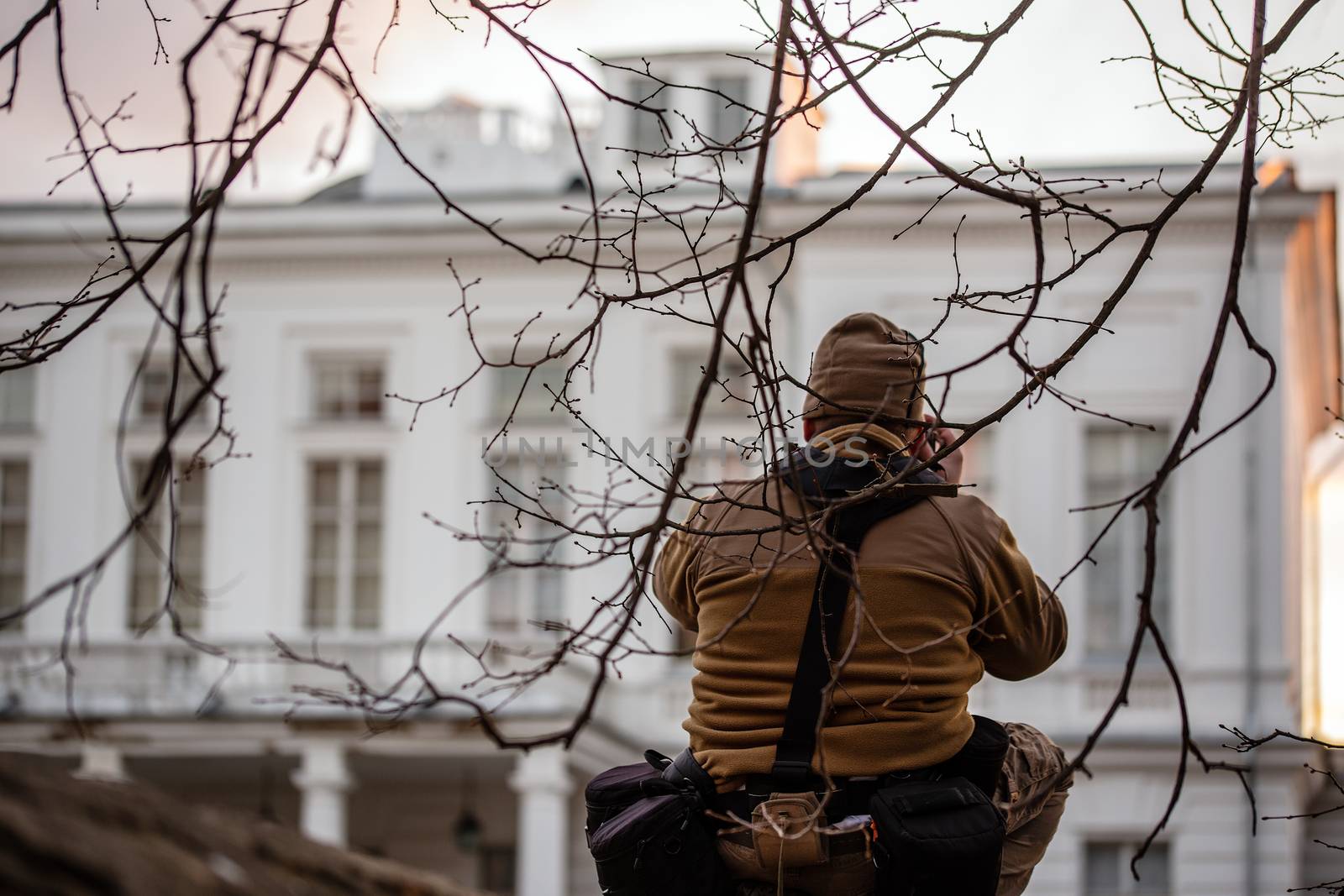 Sneaky professional photographer shooting private white building facade from fence under tiny brcnches. Pro gear weared with belt pouches and fleece coat.