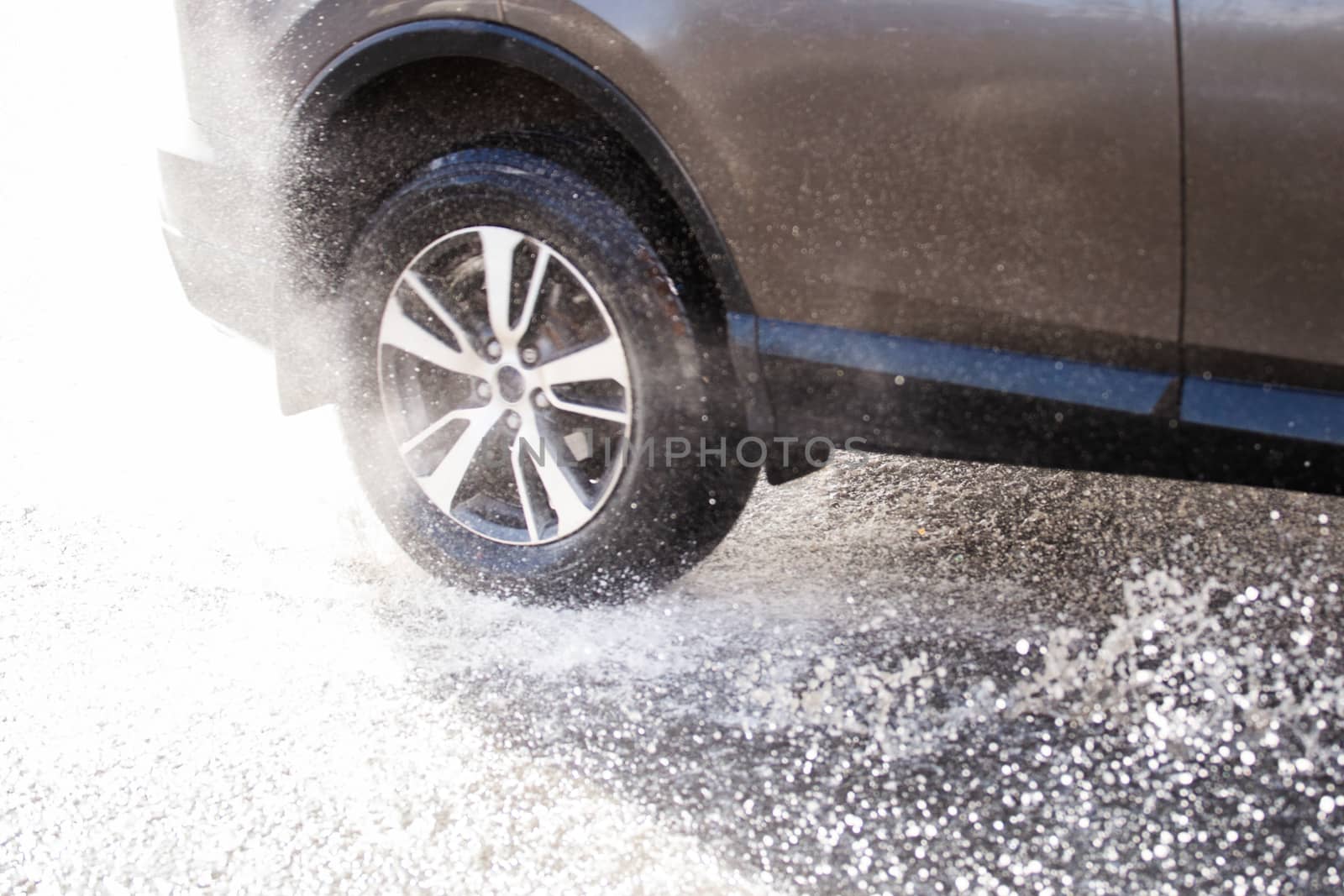 Gray car crossing puddle with water splash at daylight closeup with defocus technique and blur in counter light.