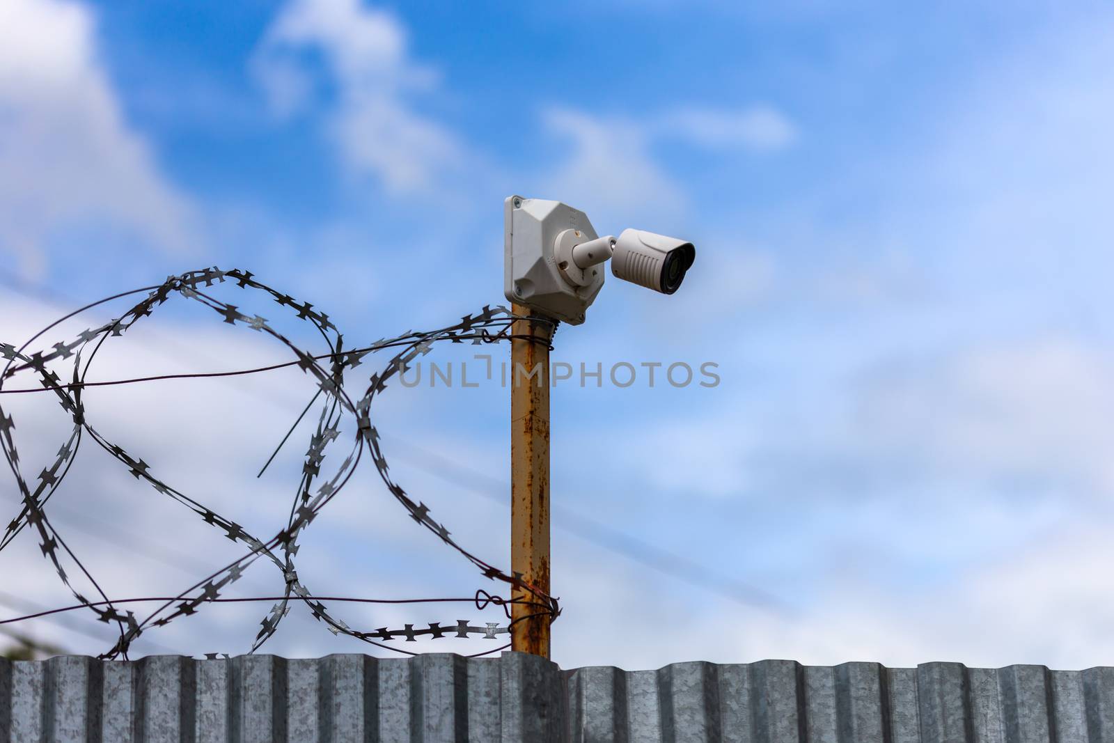 Profiled sheet fence with cheap surveillance camera and barbwire with selective focus at daylight on blue sky with clouds blurred background.
