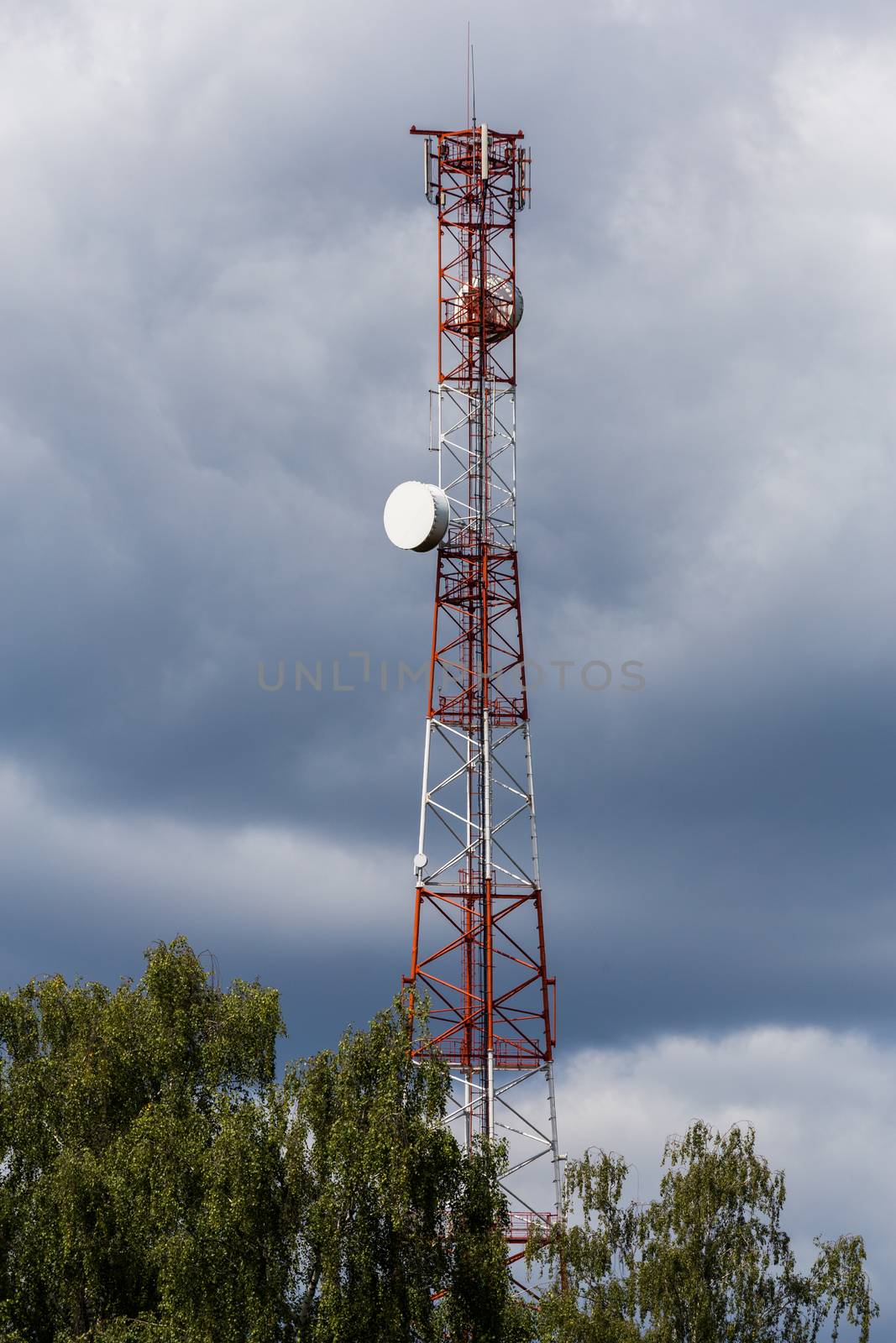 Red-white telecommunication tower with top of green tree on blue sky with clouds background,