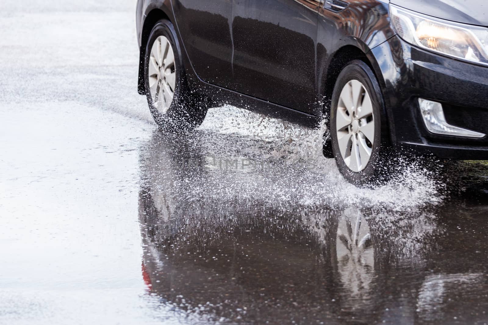 Black car crossing puddle with water splash at daylight closeup with selective focus and blur.