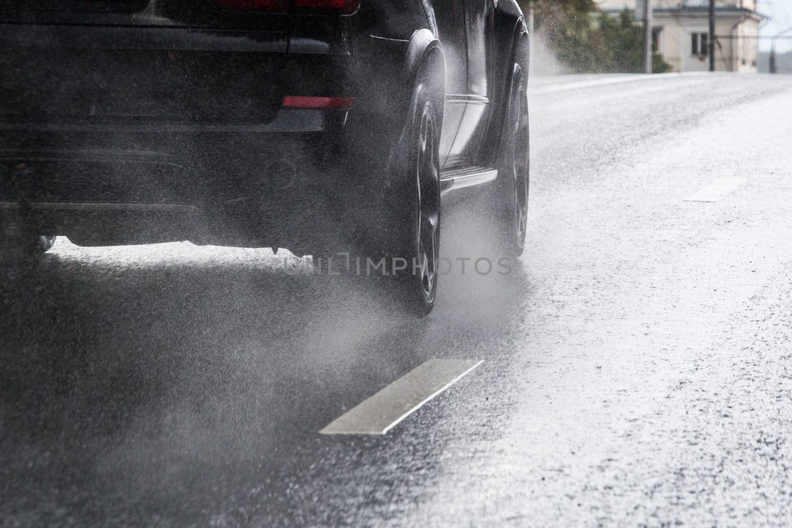 Rain water splash flow from wheels of black car moving fast in daylight city with selective focus. Tiny drops mist.