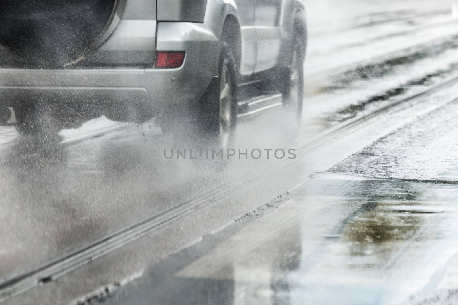 Rain water splash flow from wheels of silver metallic crossover car moving fast in daylight city with selective focus. Tiny drops mist.