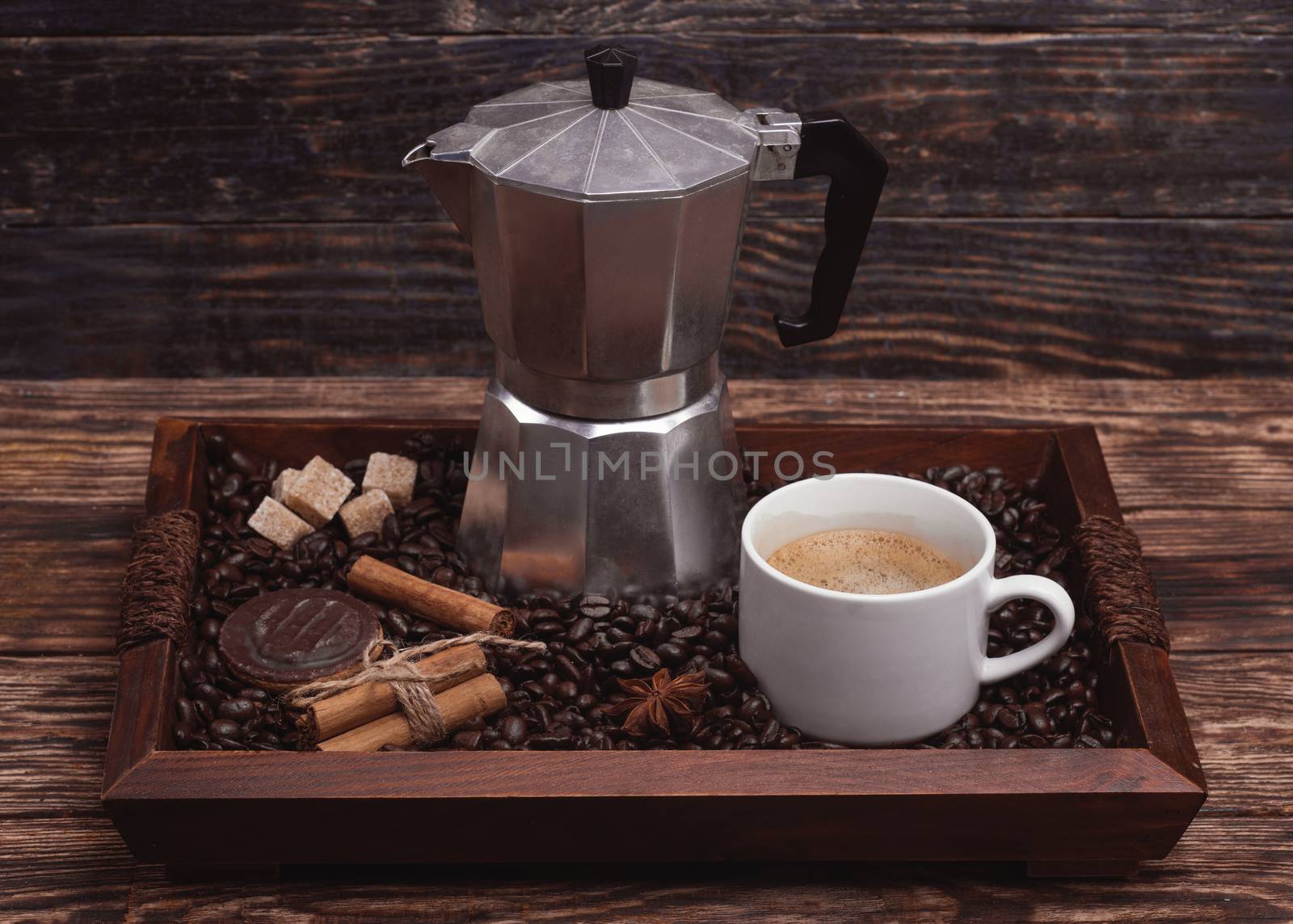 Cup of coffee, coffeepot, biscuit, cinnamon, anise, sugar, coffee beans on a wooden tray.