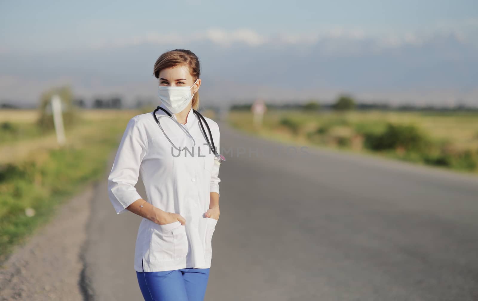 Female doctor or nurse wearing a protective face mask next to a rural road. by selinsmo