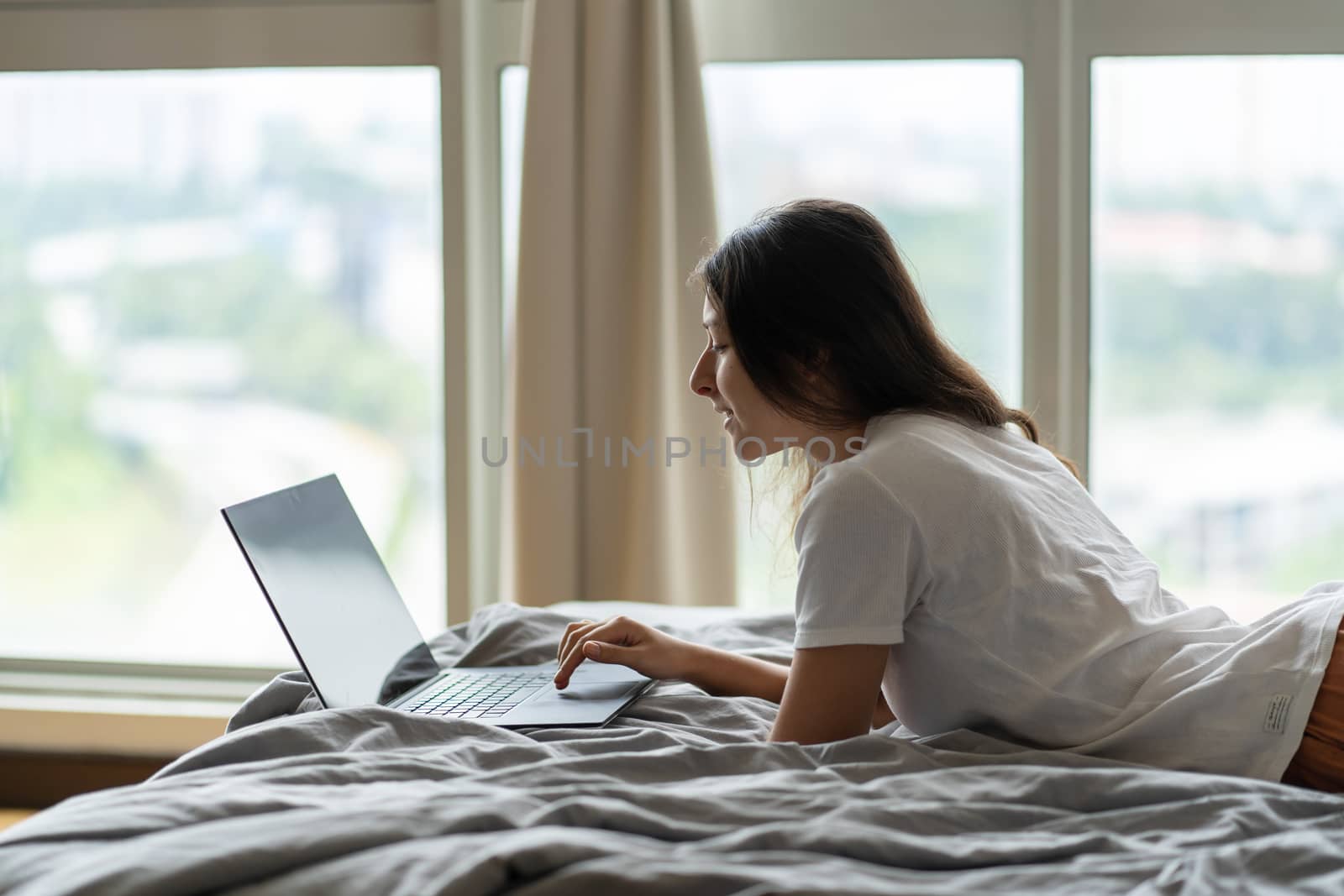 A beautiful young brunette girl is working on the laptop while lying on a bed by a panoramic window. Stylish modern interior. A cozy workplace. Shopping on the Internet