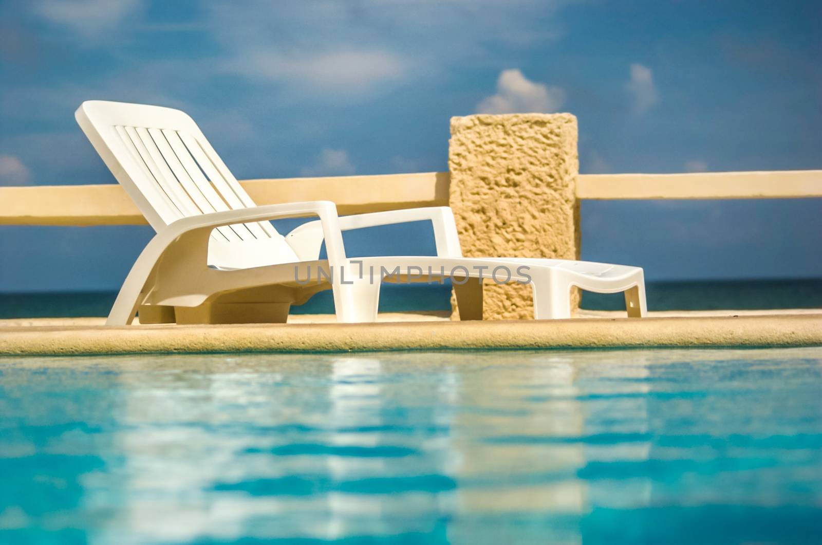 Empty white plastic sunbed next to pool with sea and sky in background