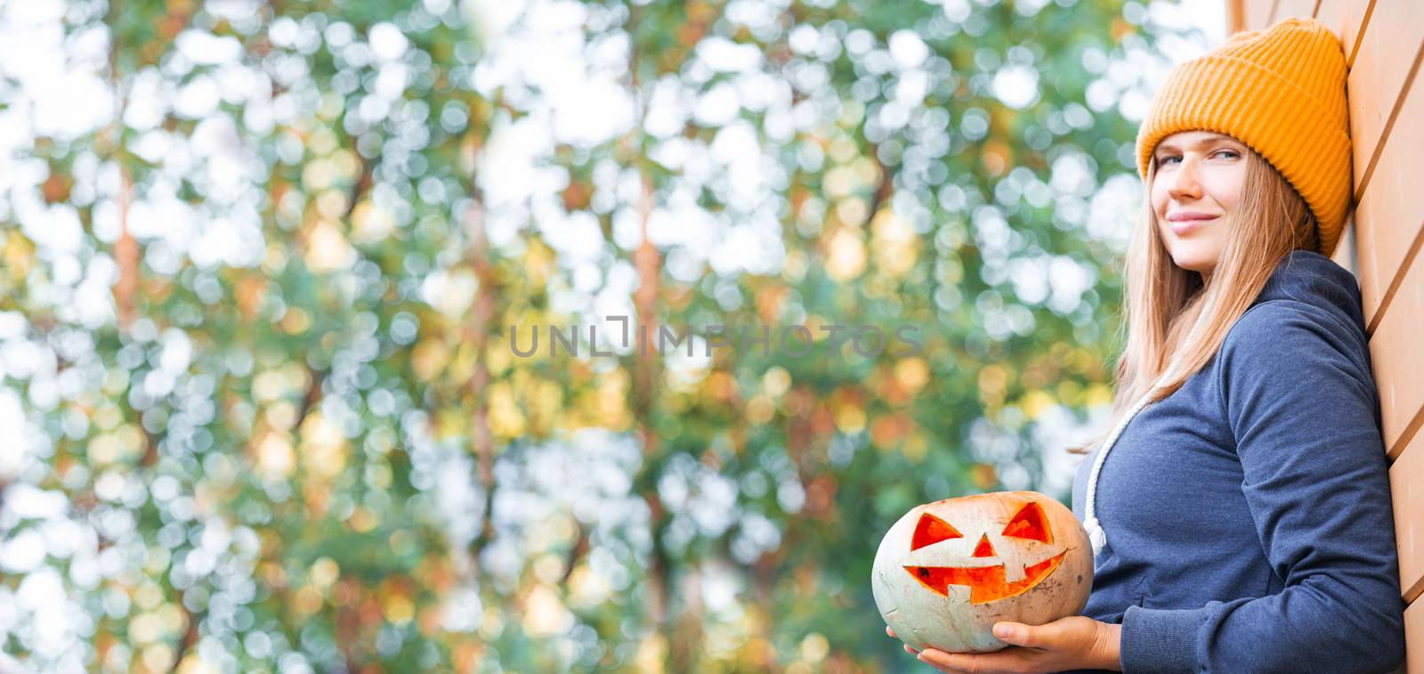Woman in casual clothes and hat holding Halloween pumpkin over autumn nature background with copy space for text