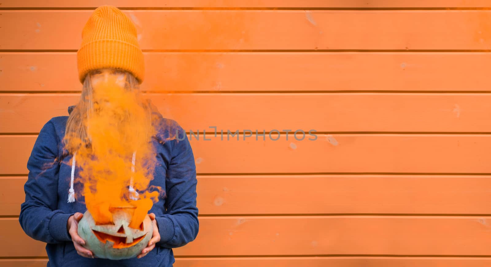 Woman in casual clothes and beanie holding Halloween pumpkin over orange wall background with copy space for text