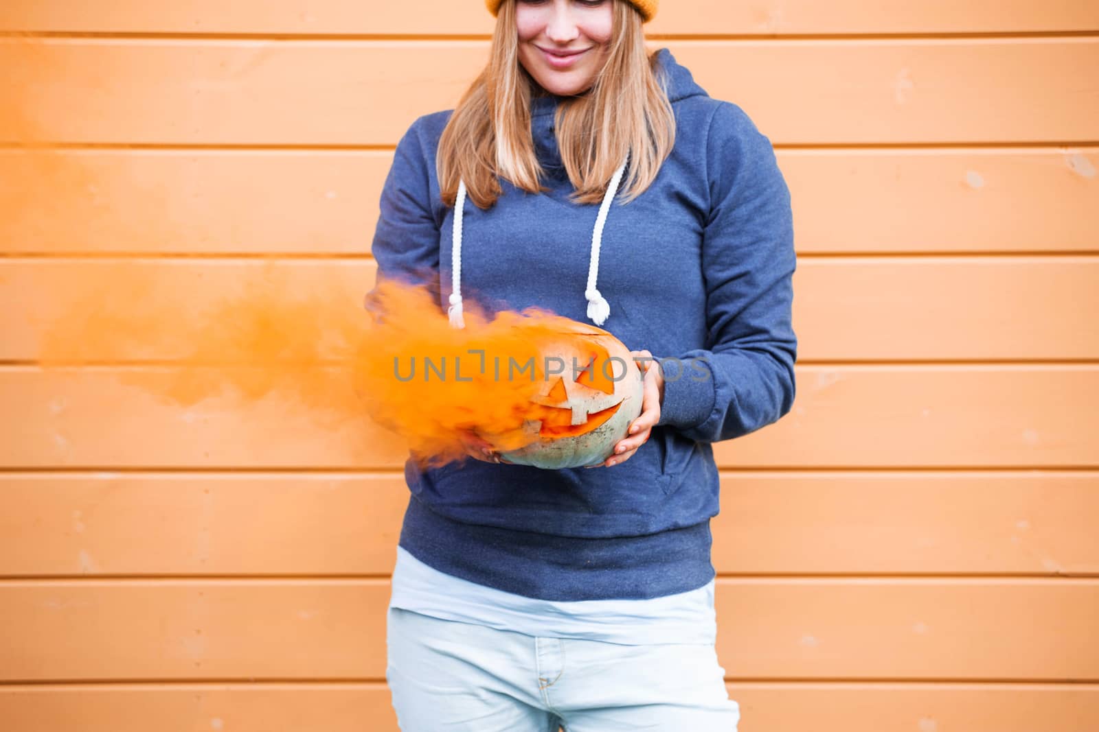 Woman in casual clothes and beanie holding Halloween pumpkin over orange wall background with copy space for text